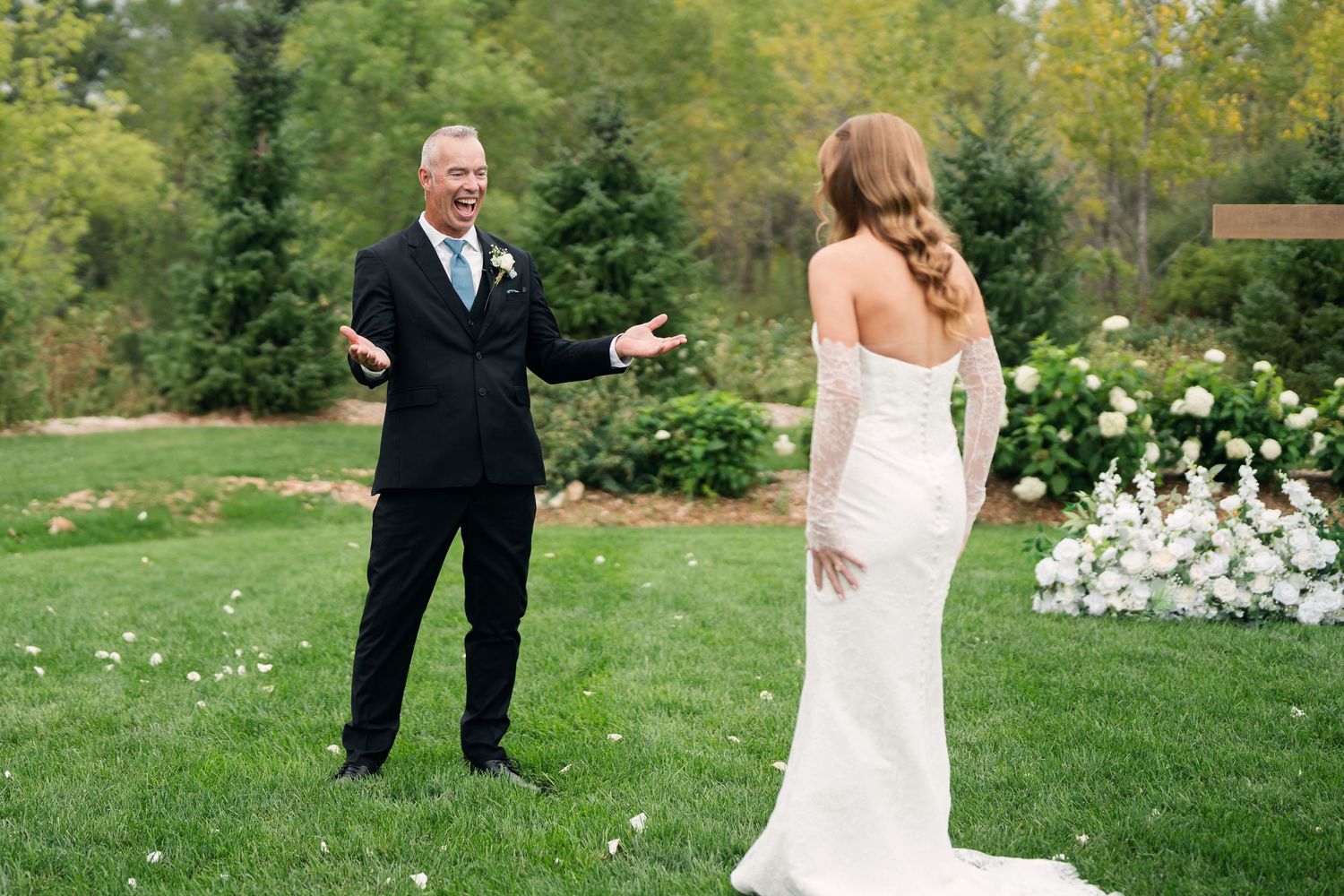 Father reacts with joy seeing bride in white wedding gown during outdoor first look on lush green lawn.