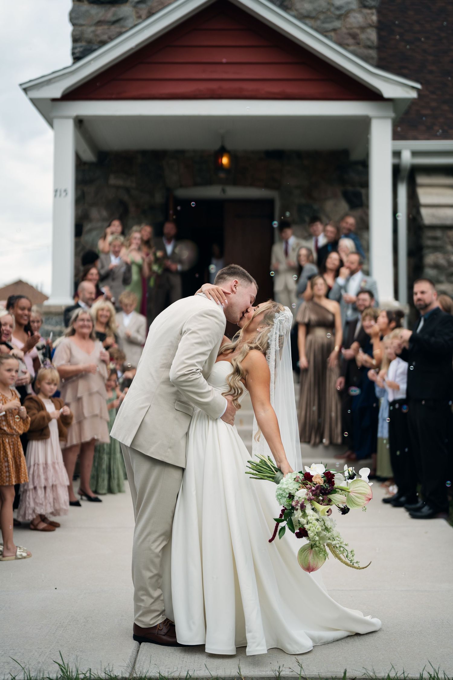 Bride and groom share first kiss outside stone church surrounded by cheering guests blowing bubbles at outdoor wedding ceremony.