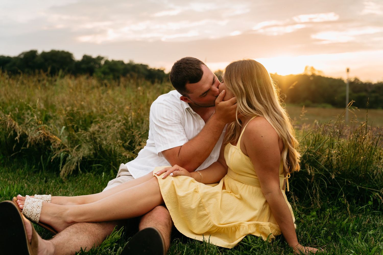 A couple shares an intimate moment in a golden meadow at sunset while sitting in tall grass.