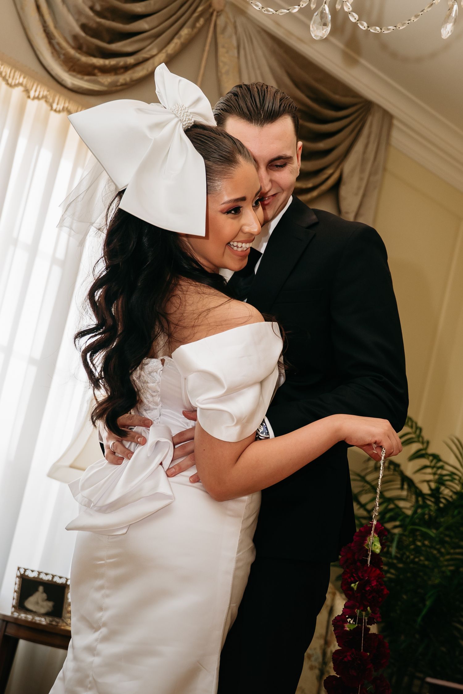 Bride in white off-shoulder gown with oversized bow headpiece embraces groom in black suit during wedding ceremony.