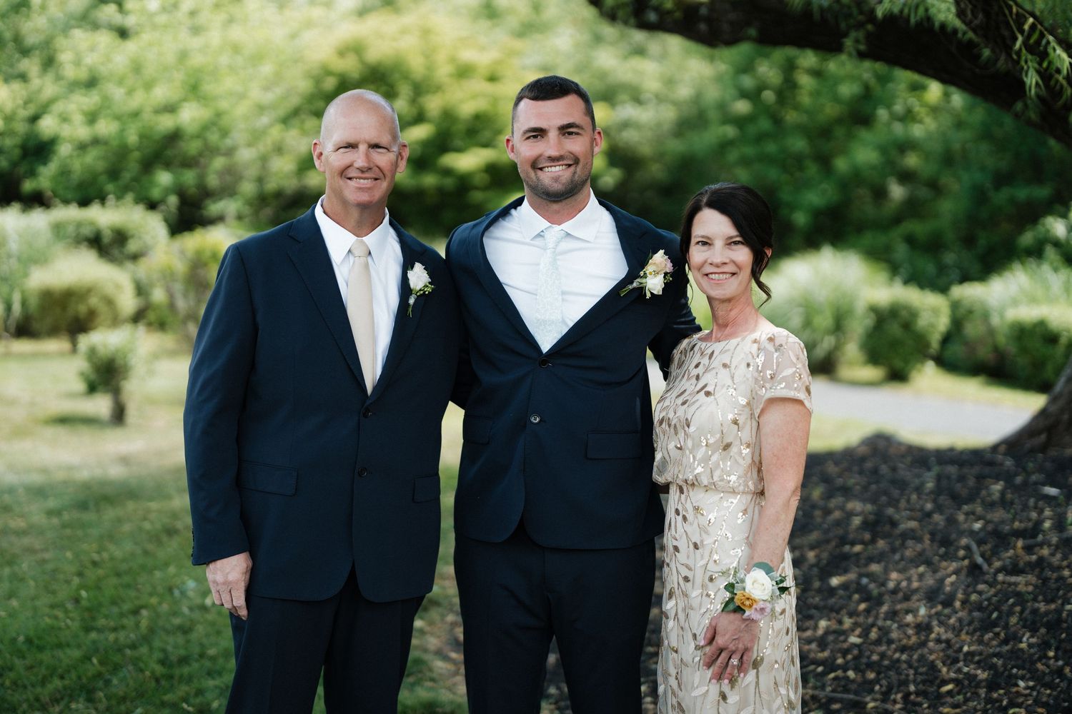 Groom posing with parents in formal attire for family portrait in lush garden setting.