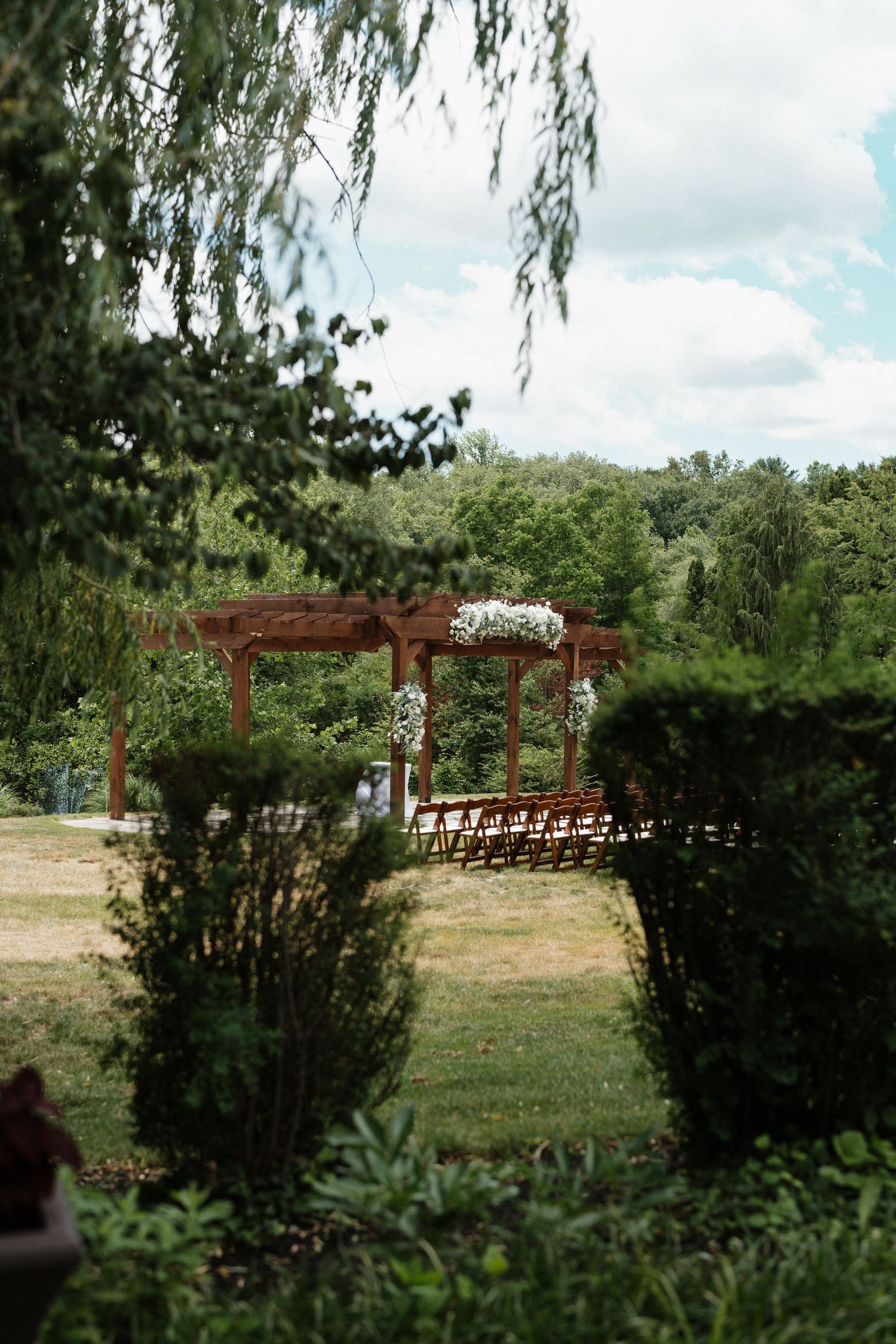 Outdoor wedding ceremony setup with wooden pergola decorated with white flowers, surrounded by lush green trees and manicured shrubs.