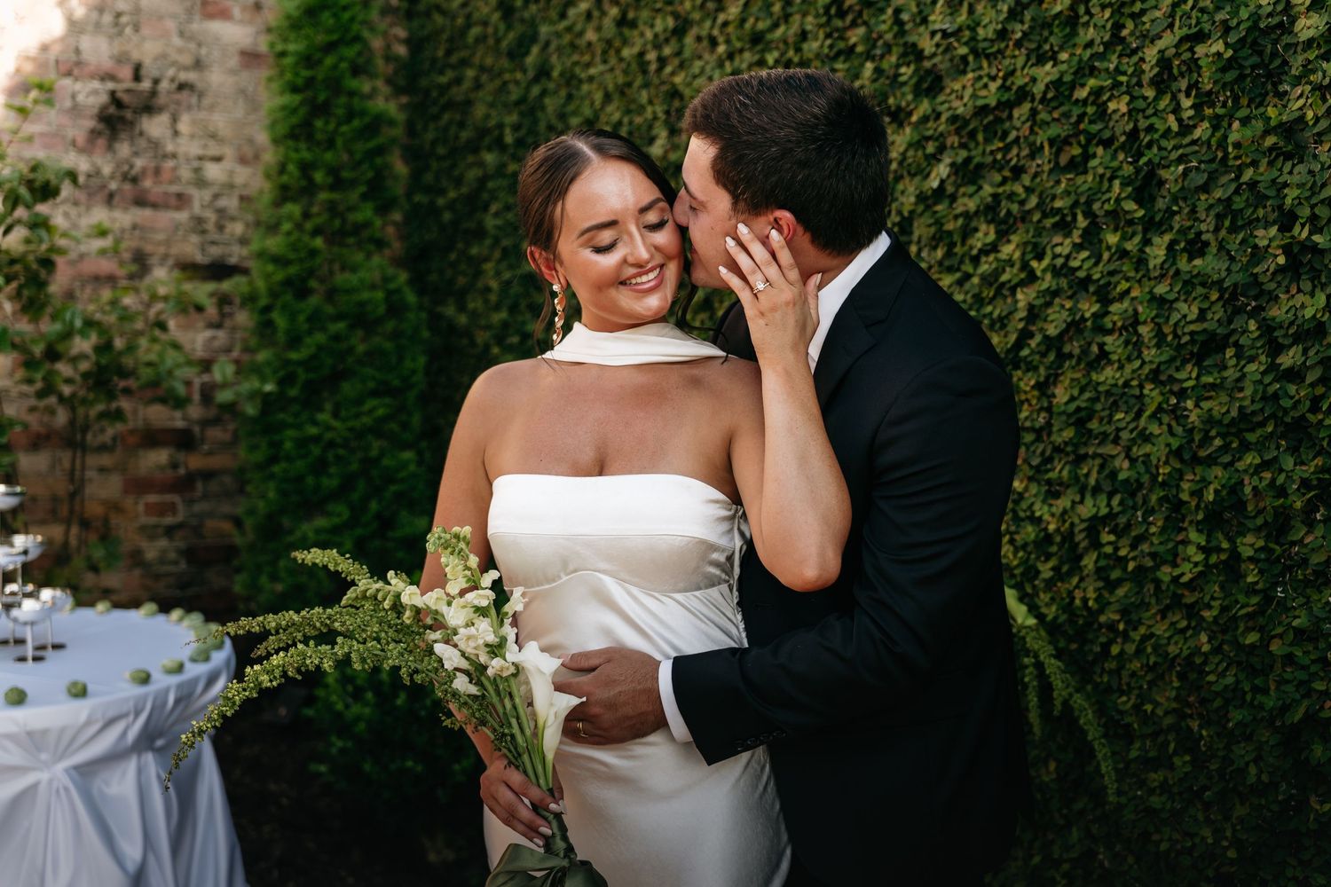 A couple shares a romantic moment in front of a lush green hedge during their outdoor wedding celebration.