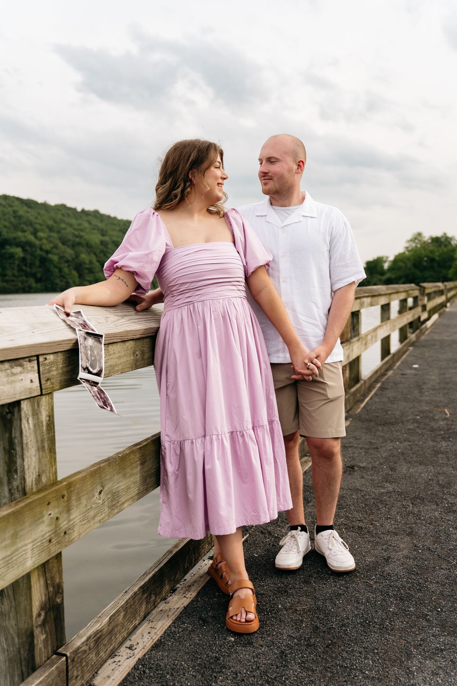 A couple in coordinating pink and white outfits share romantic moments on a wooden pier overlooking a tranquil lake.
