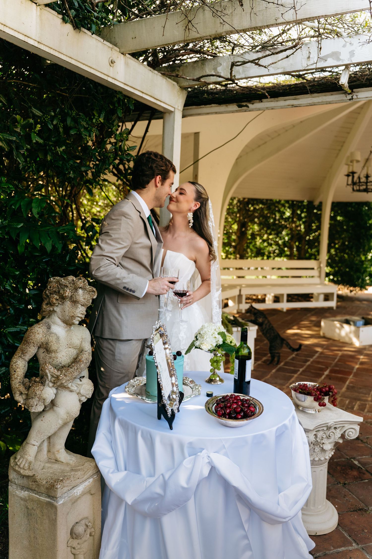 A romantic wedding couple shares intimate moments at an elegant outdoor venue with white tablecloths and stone statues.