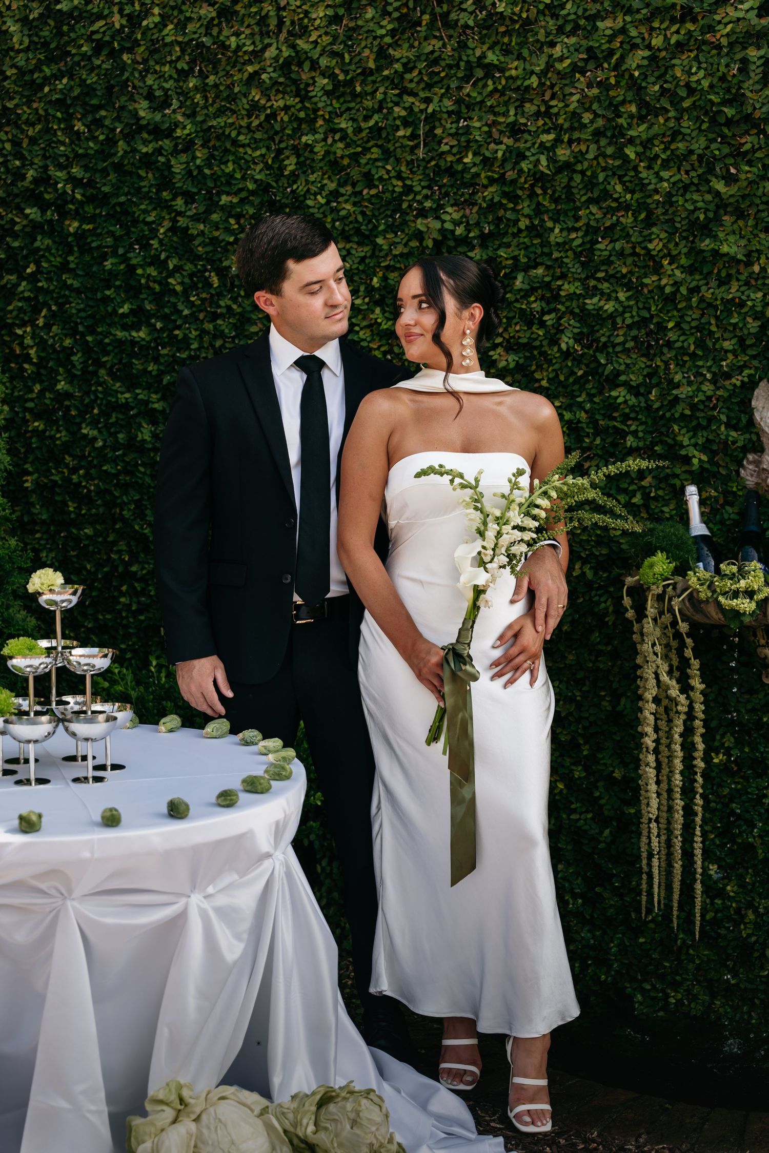 Wedding couple poses against green hedge backdrop with elegant white table settings and champagne glasses.