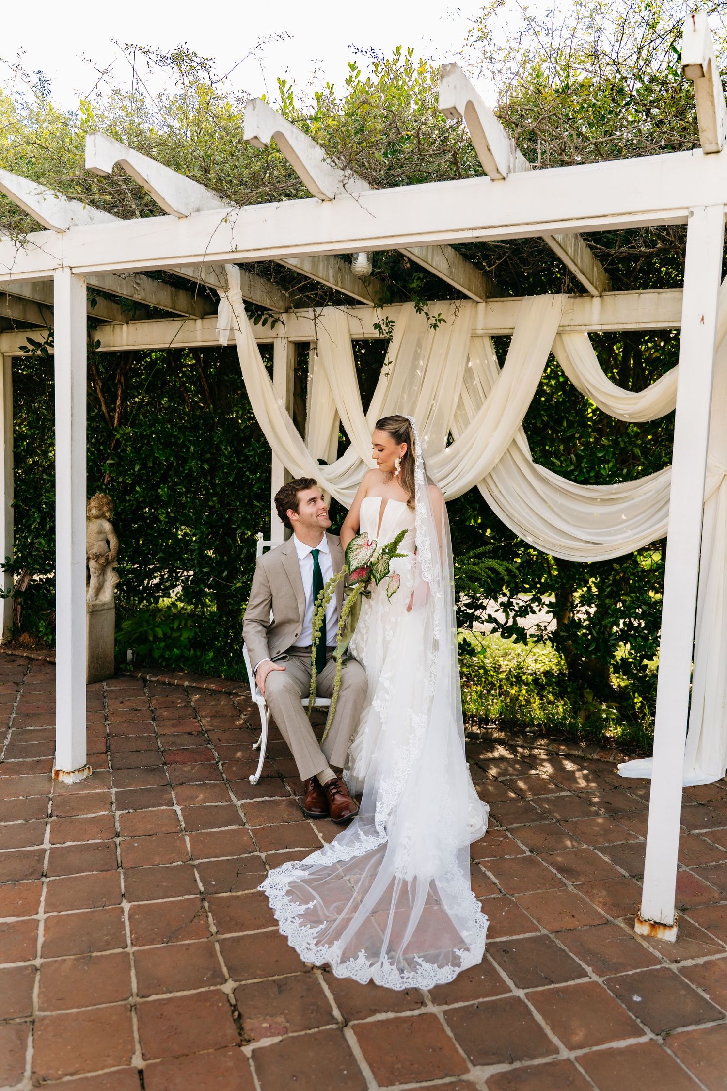 Bride and groom share romantic moment under draped pergola, bride in lace gown with long train.