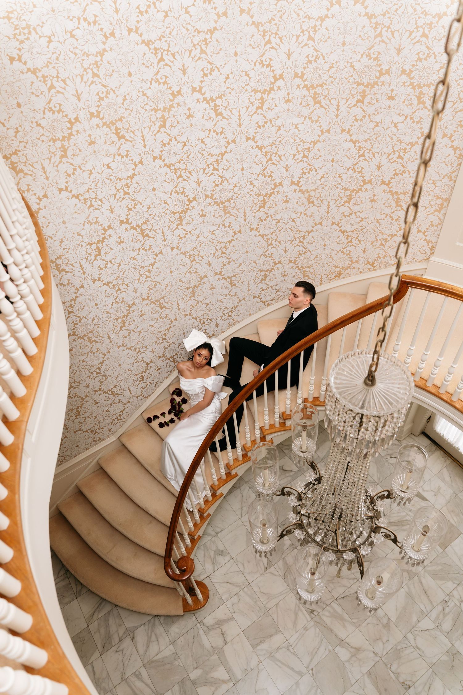 Bride and groom sitting on elegant curved staircase with crystal chandelier in ornate wallpapered venue.
