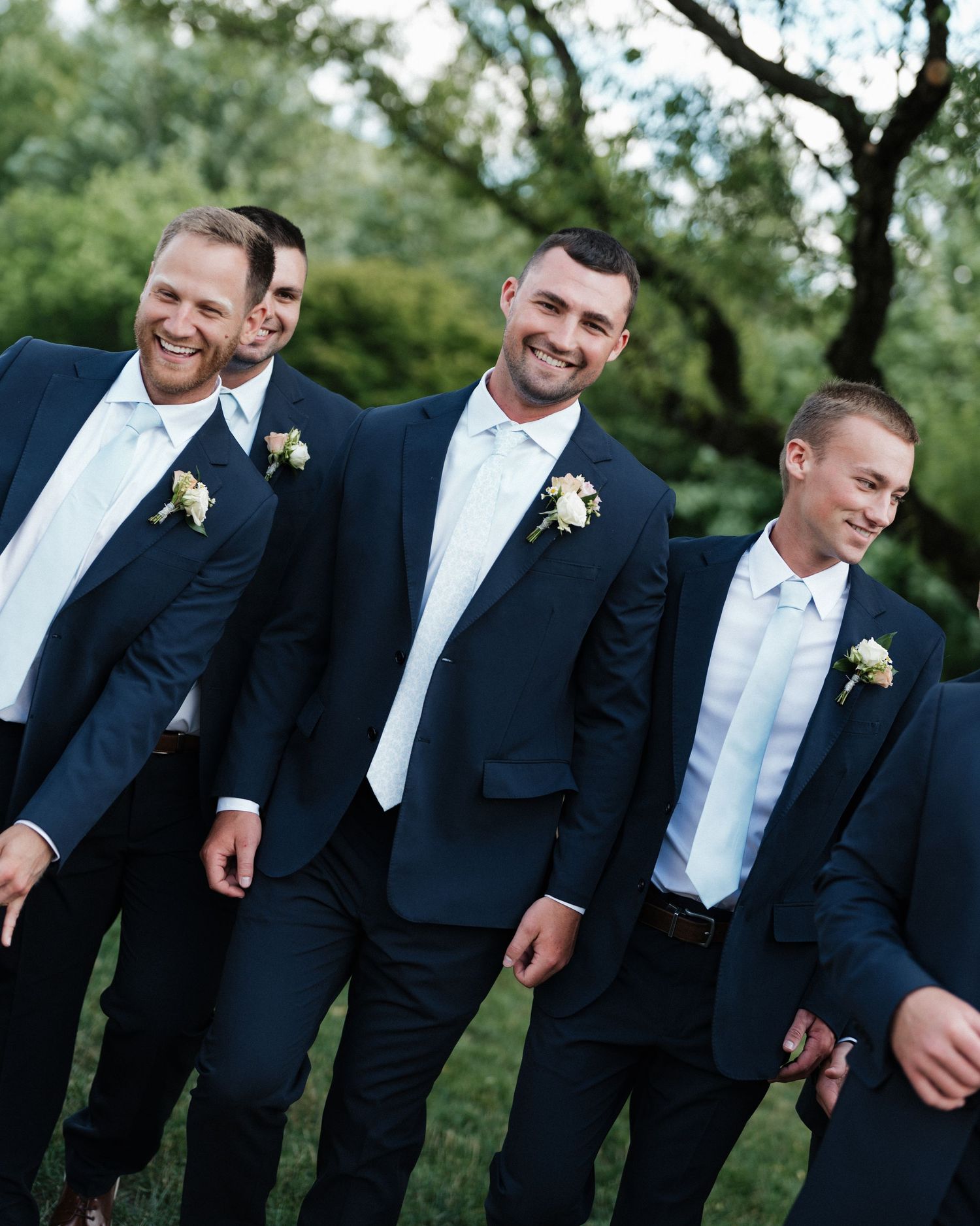 Groom and groomsmen laughing in navy suits with boutonnieres outdoors at wedding celebration.