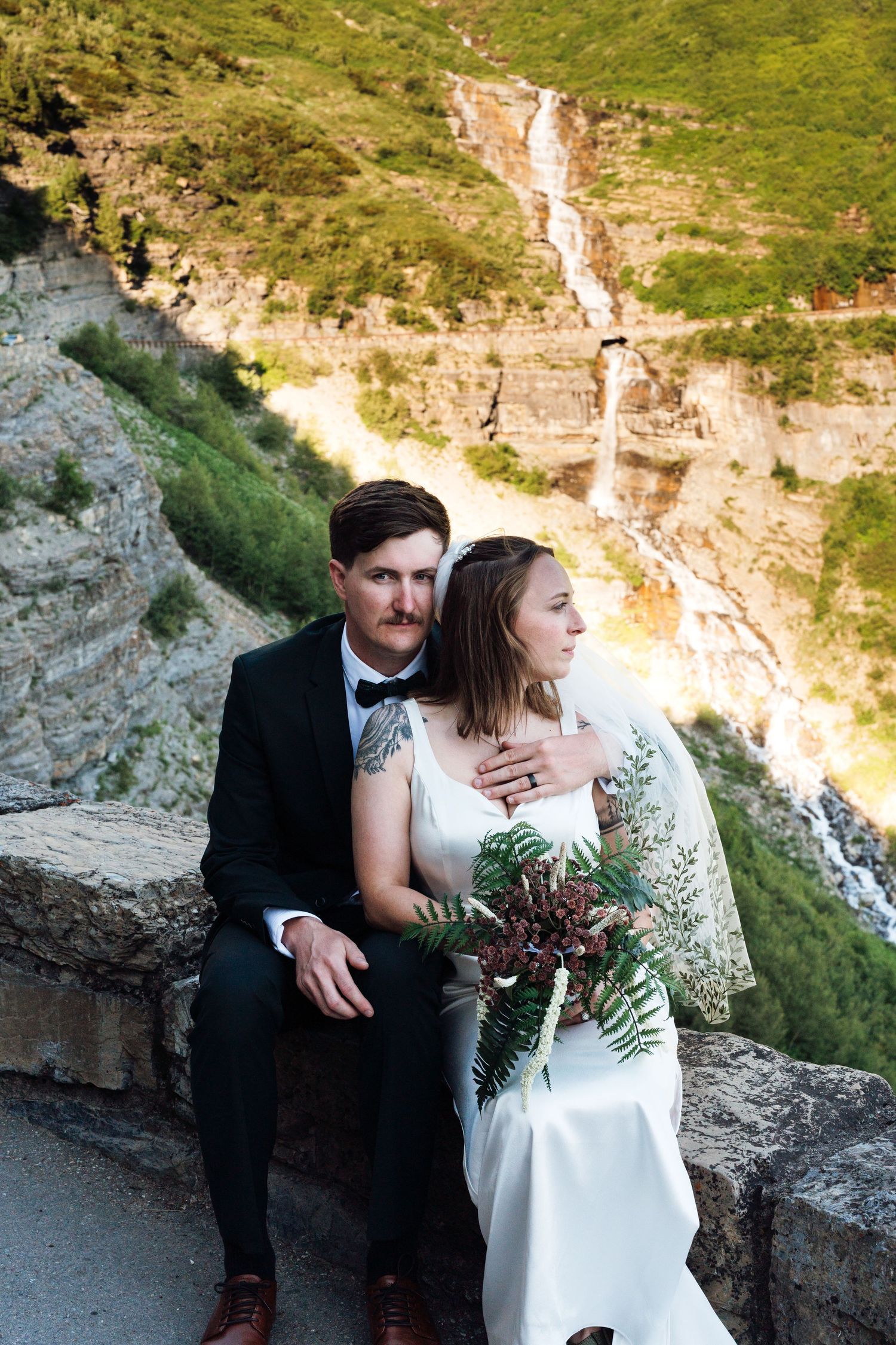 Bride and groom sit on stone wall with waterfall and green canyon backdrop, bride holding fern bouquet.