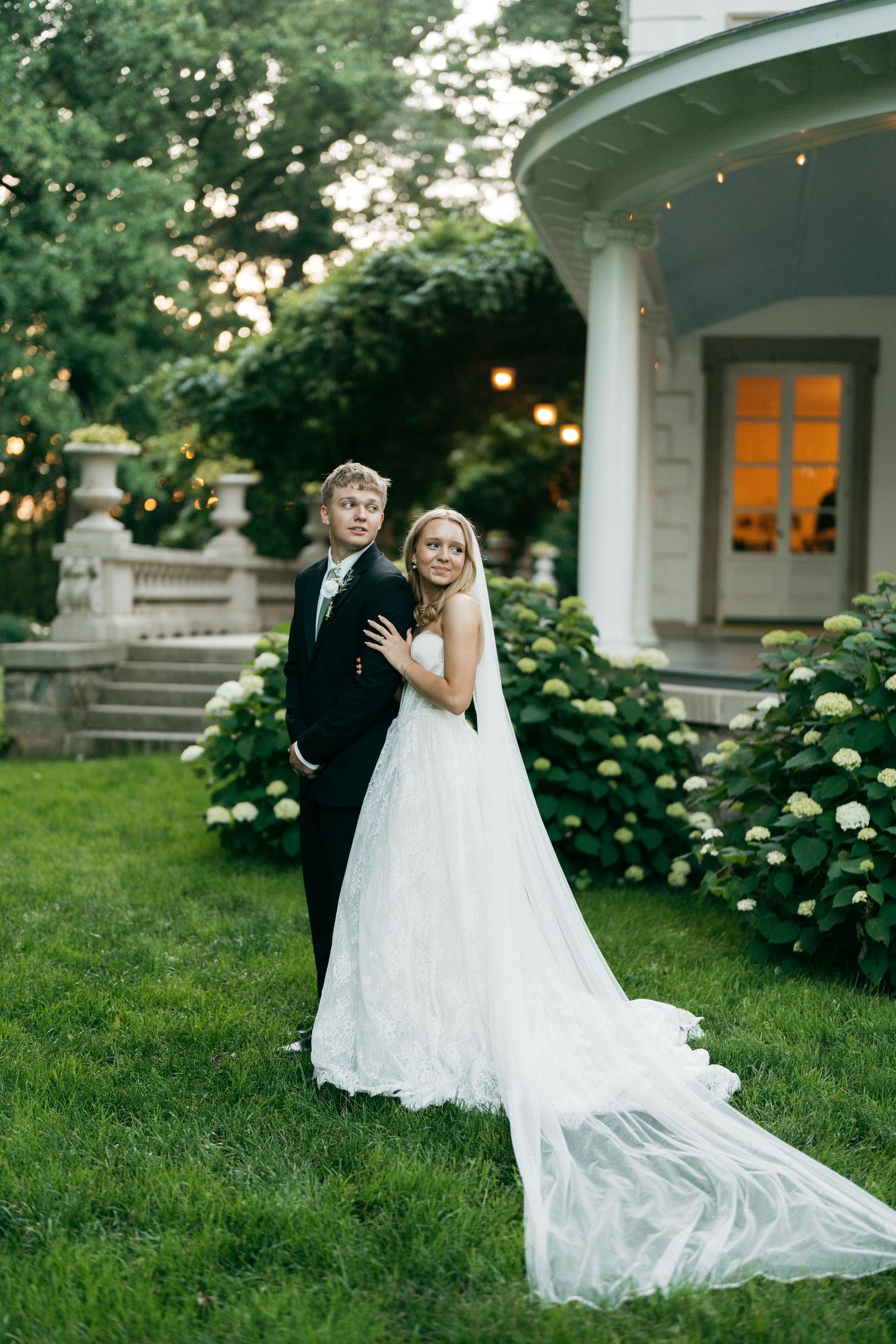 Bride in white gown with train and groom in black suit pose on green lawn beside elegant mansion with hydrangeas.