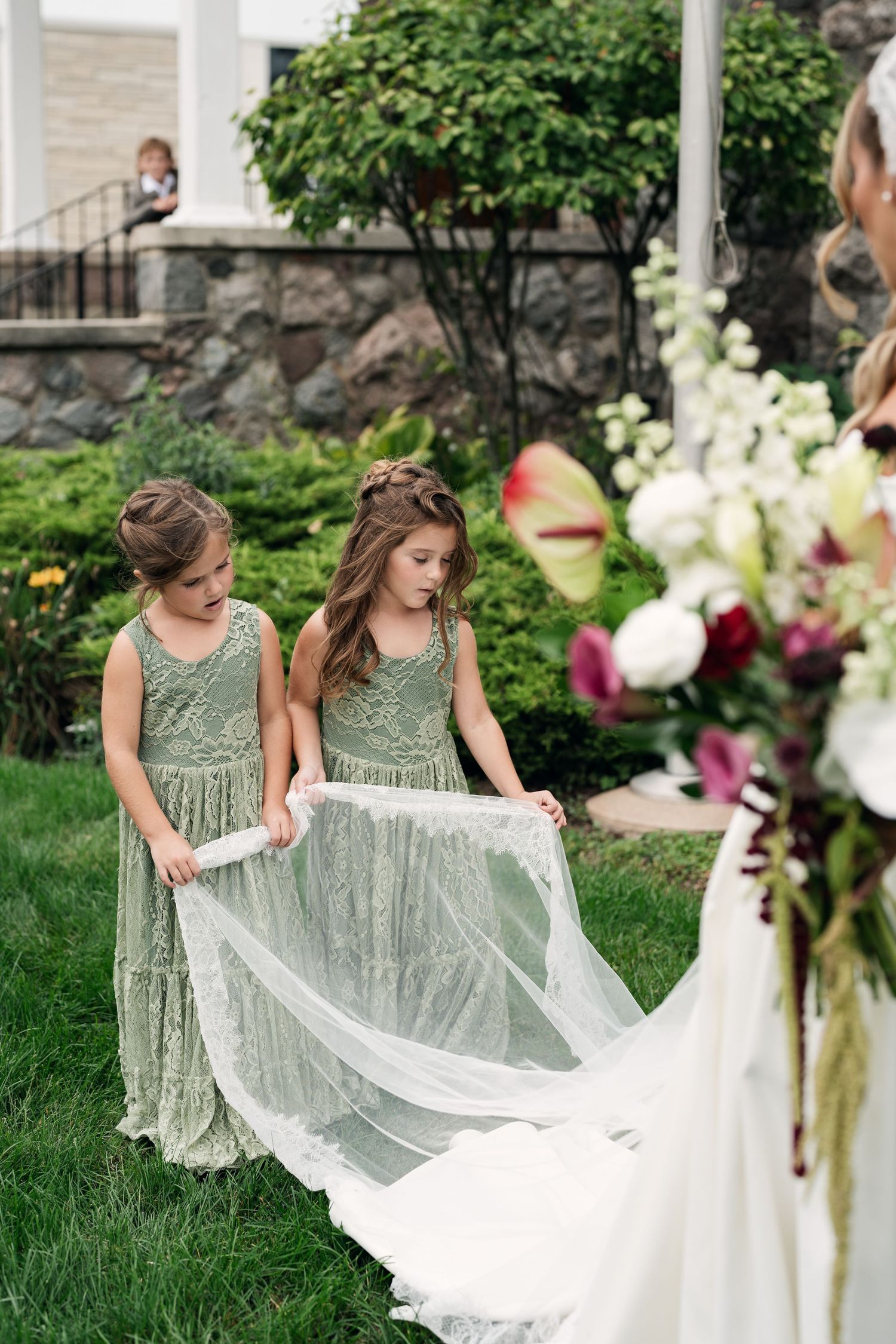 Two flower girls in sage green dresses hold a bride's white veil train outdoors at a garden wedding ceremony.