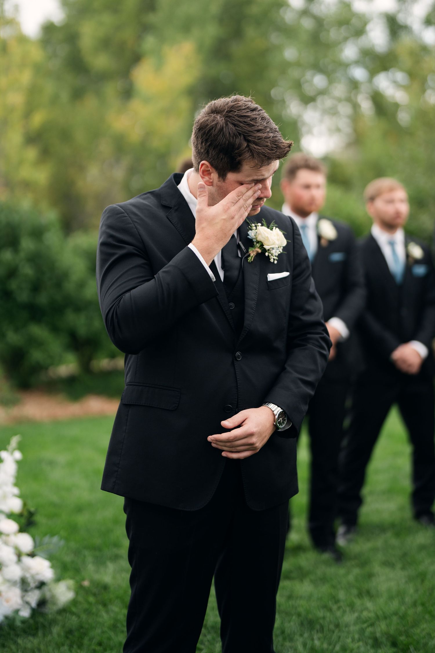 Emotional groom crying while waiting at the altar during outdoor wedding ceremony in black suit with boutonniere.