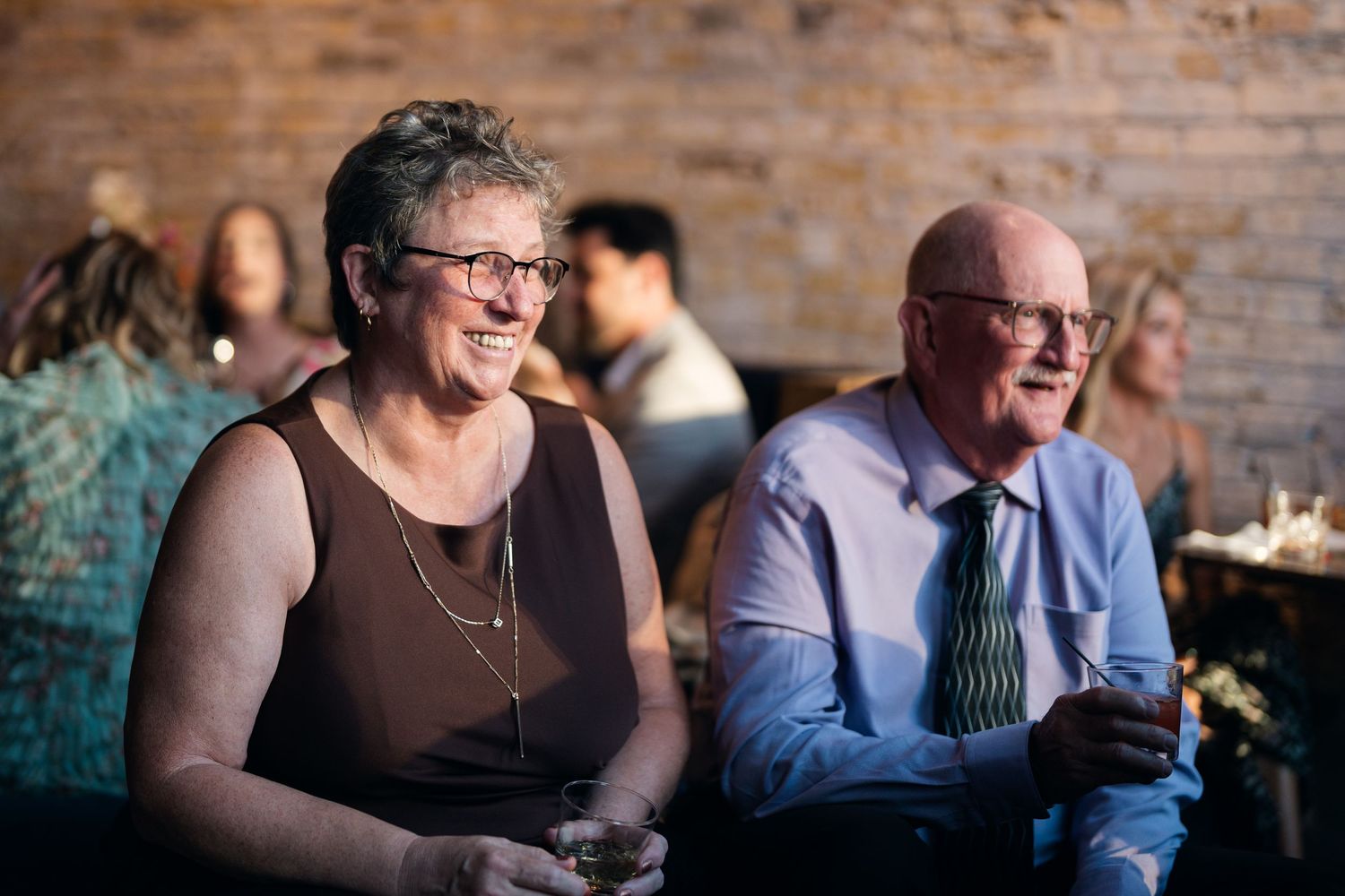 Smiling older couple enjoying drinks at a wedding reception in a warmly lit brick venue.