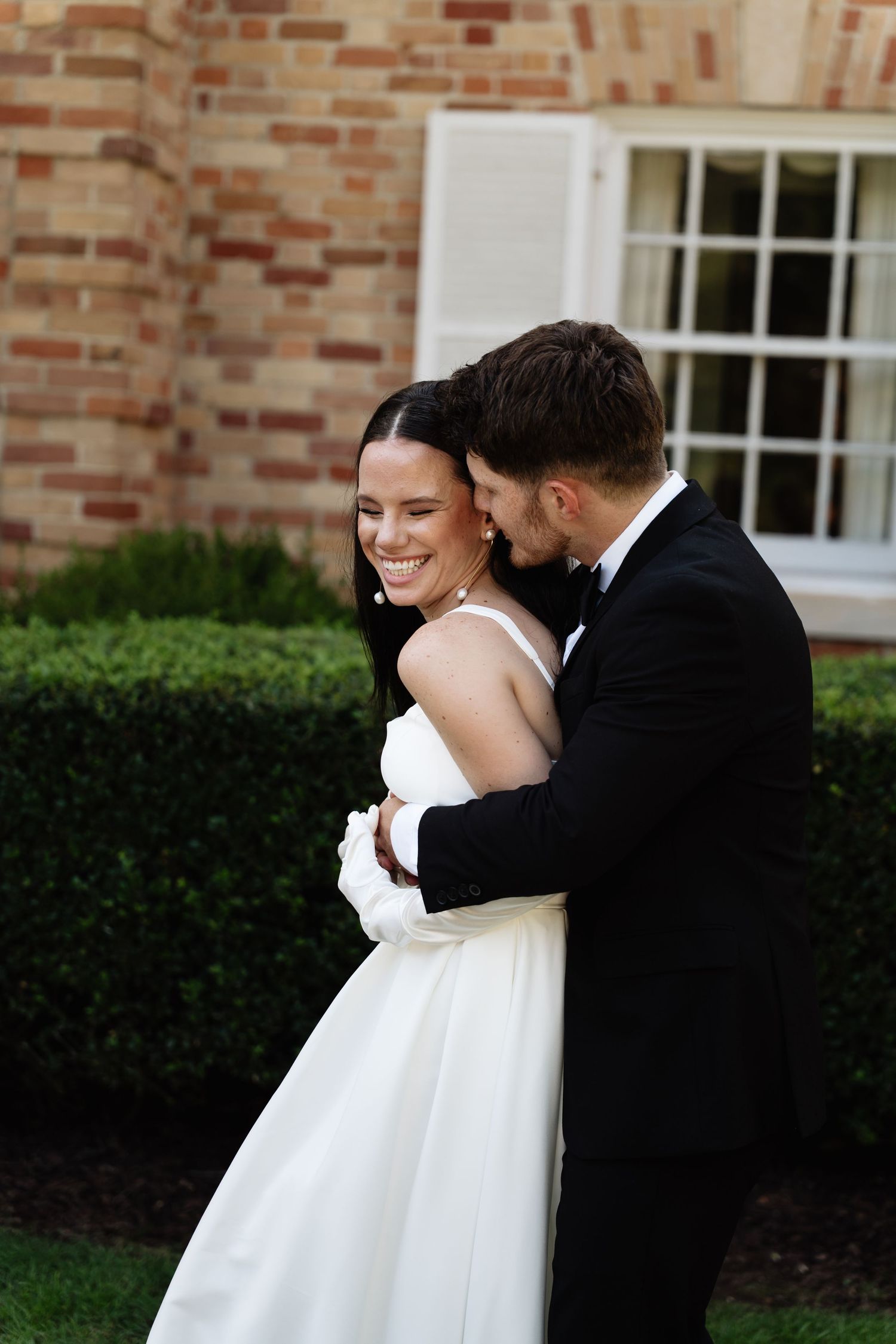 A bride and groom share an intimate moment outdoors against a brick wall and white window backdrop.