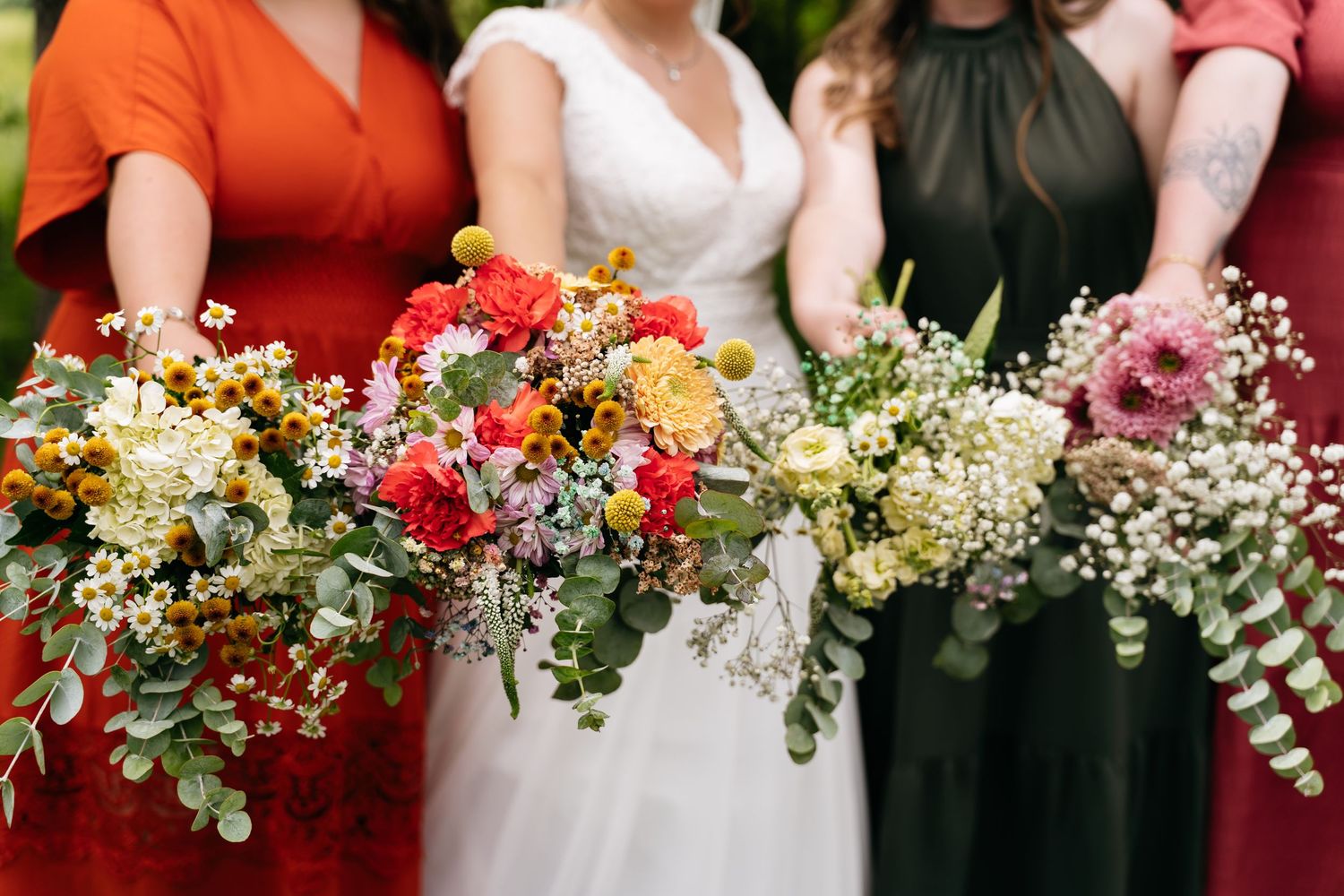 Colorful wedding bouquets with red, yellow and white flowers displayed in a row against bridesmaid dresses.