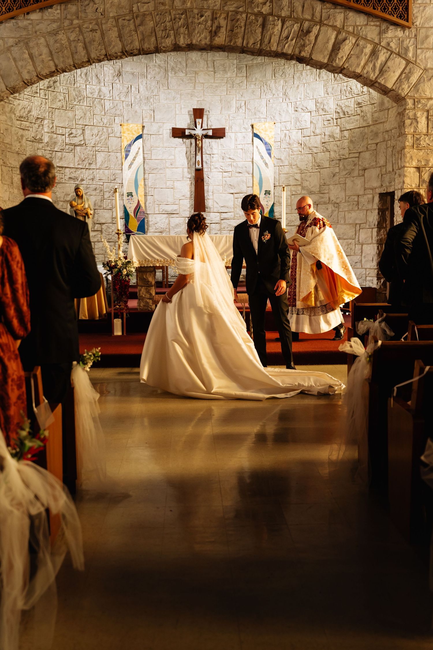 A traditional wedding ceremony takes place in a stone-walled church with people gathered around a cross-adorned altar.