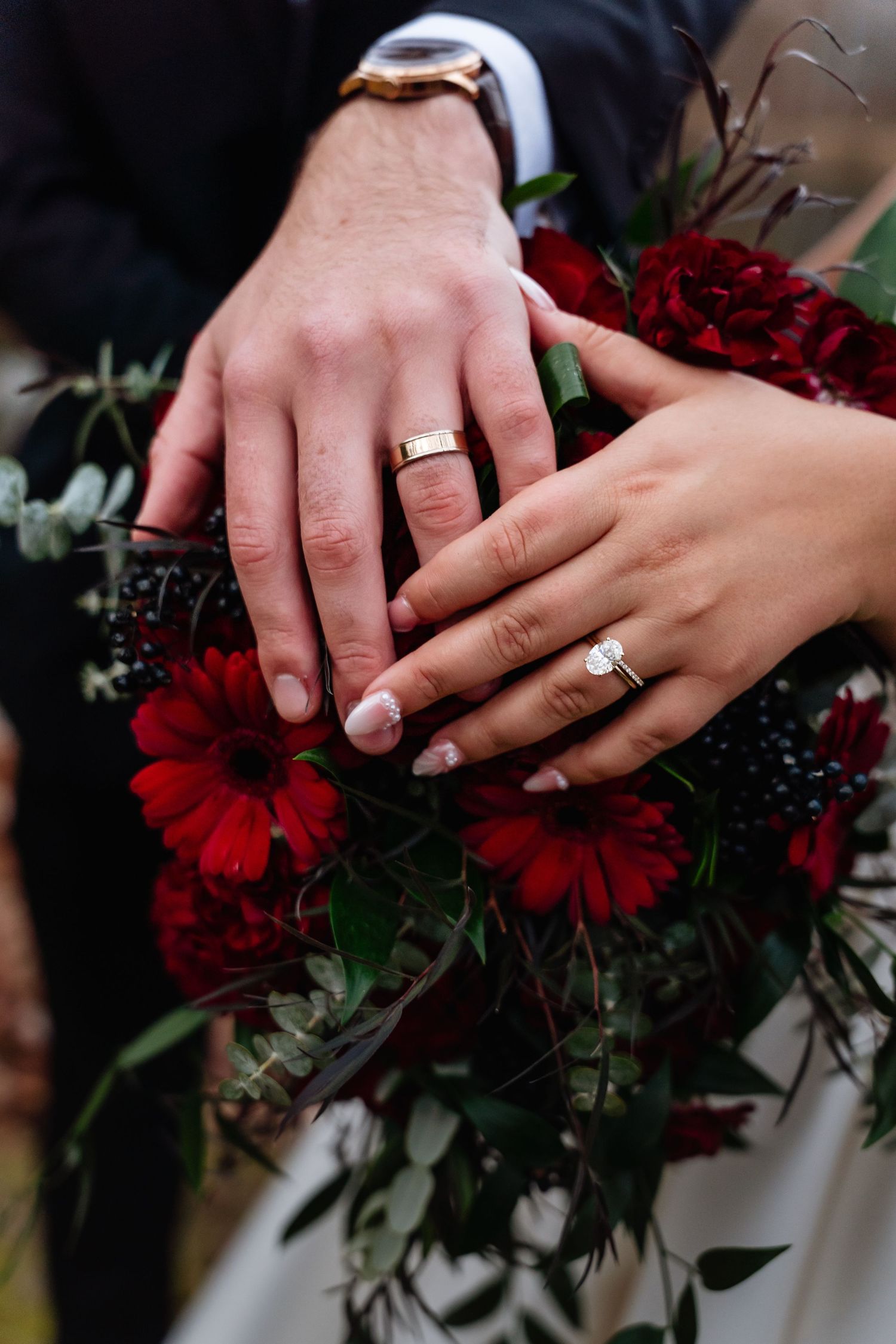 Newlyweds displaying wedding rings on a lush red floral bouquet with dark flowers and greenery.