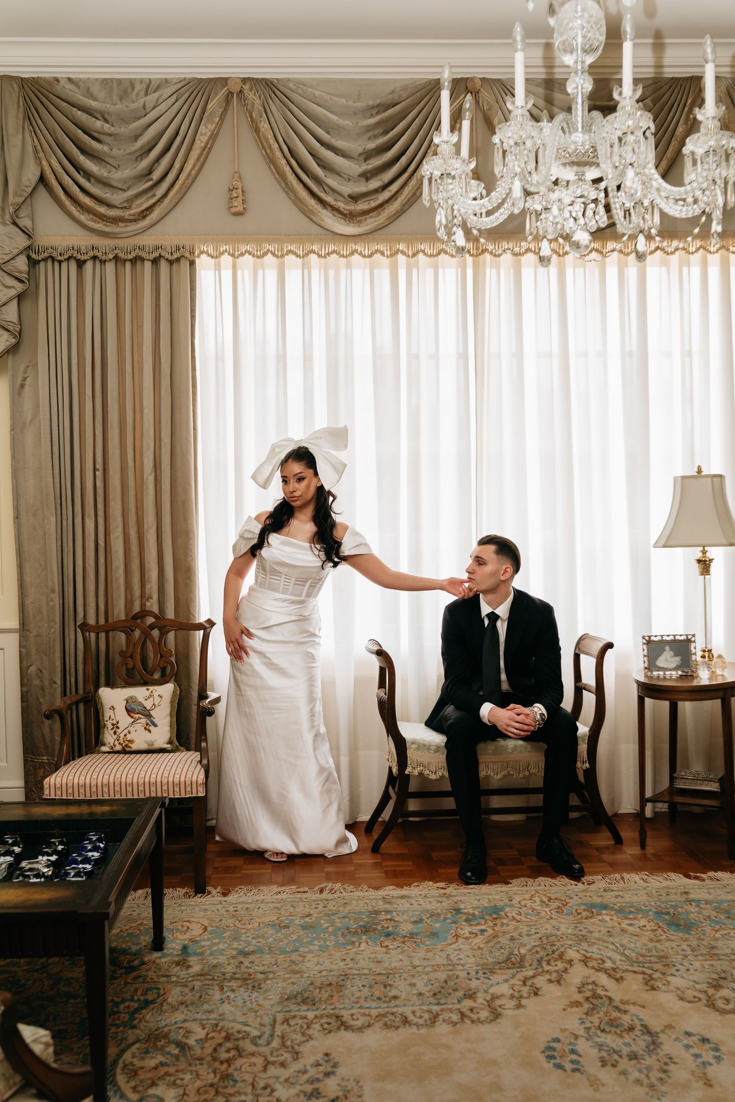Bride in white gown with bow touches groom's face in elegant room with chandelier and draped curtains.