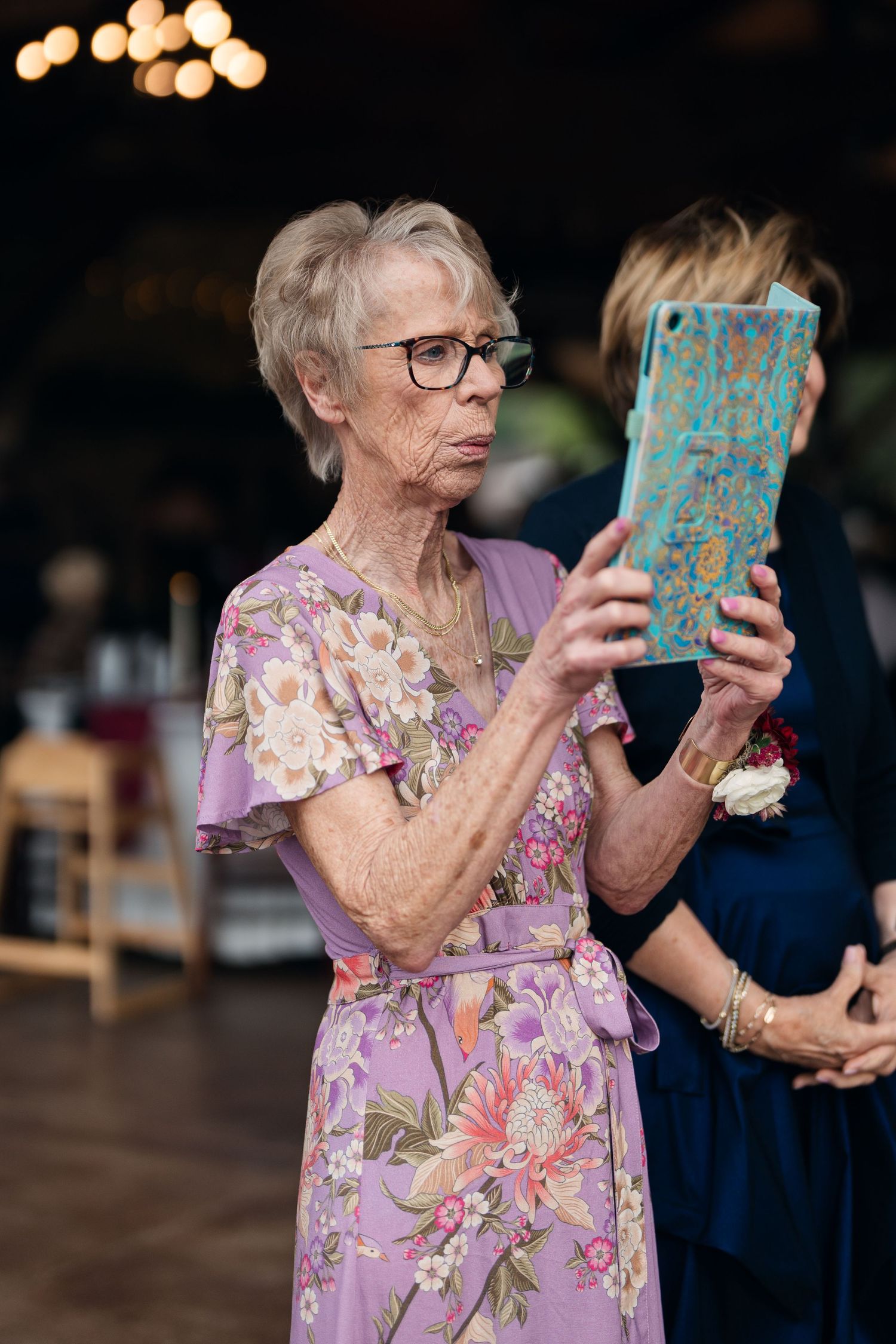 Elderly woman in floral purple dress using a teal tablet to photograph at an indoor wedding reception.