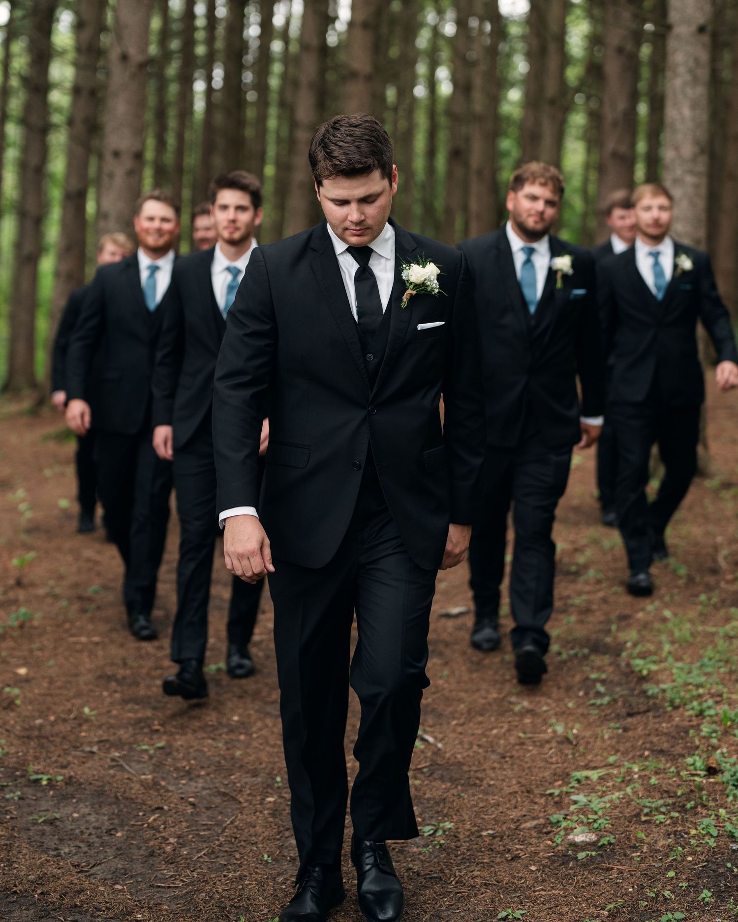 Groom leads groomsmen in black suits through forest path on wedding day.