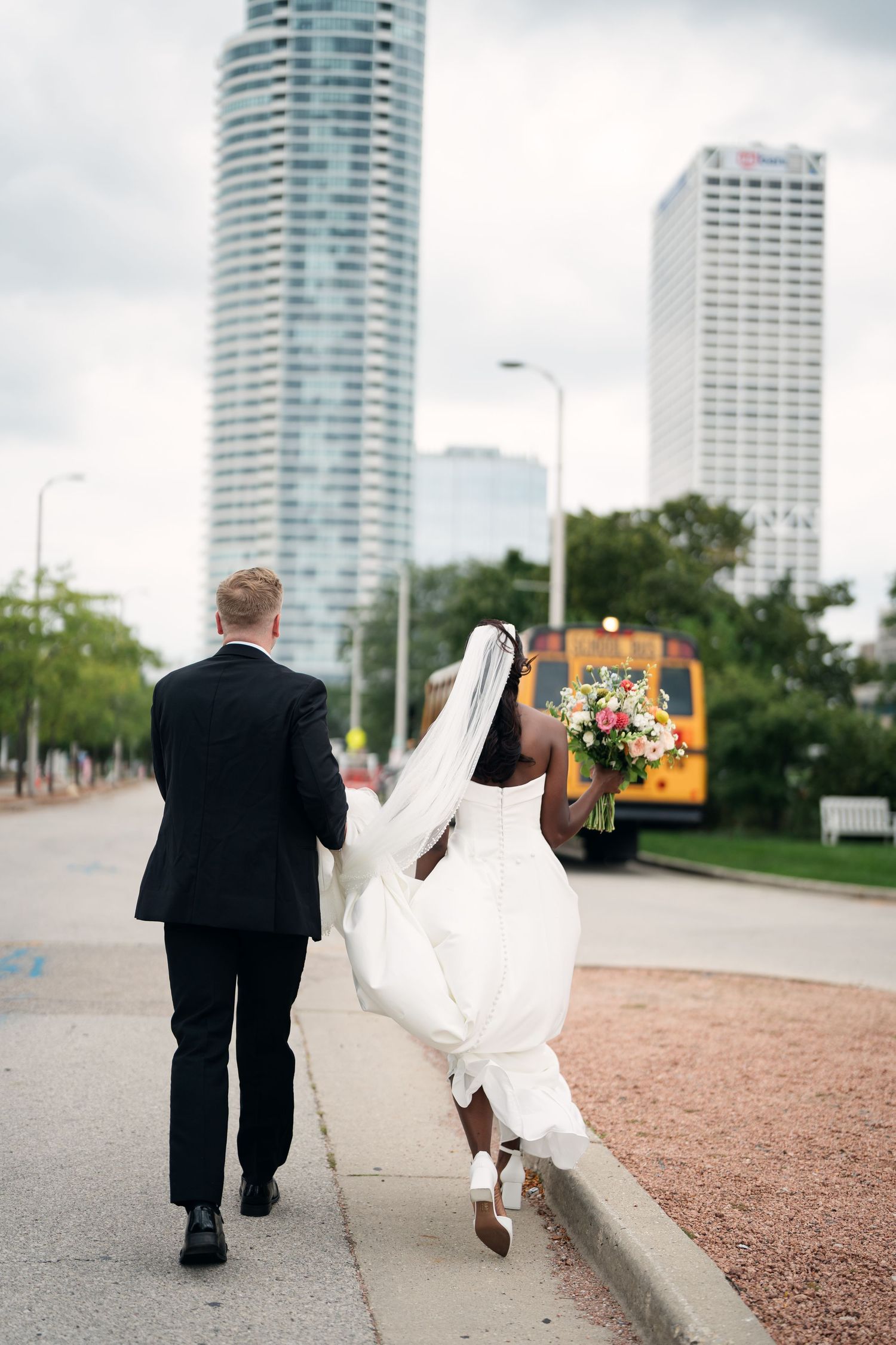 Interracial couple in wedding attire walking hand-in-hand on city sidewalk, bride holding colorful bouquet with skyscrapers behind.