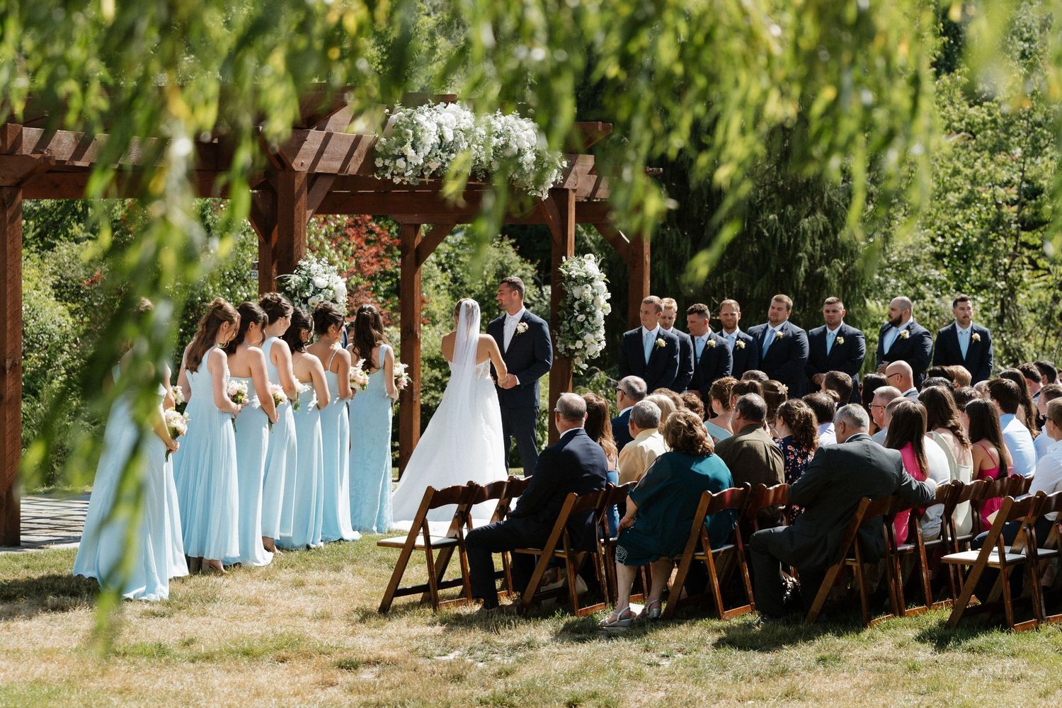 Outdoor wedding ceremony with bridesmaids in light blue dresses, groomsmen, and guests seated under a floral arbor.