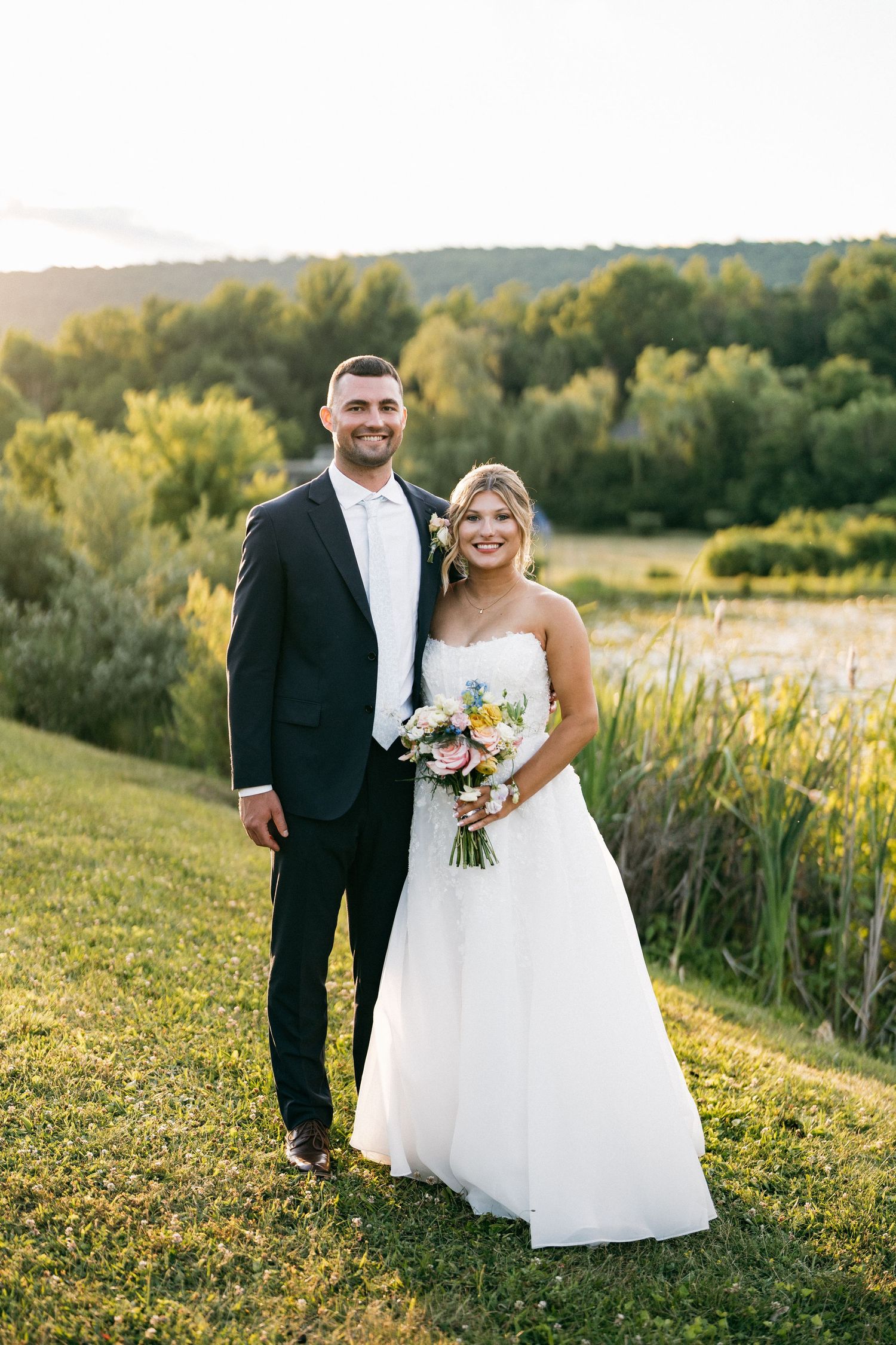 Smiling bride and groom pose outdoors at golden hour near a pond with lush greenery, bride holding a colorful bouquet.