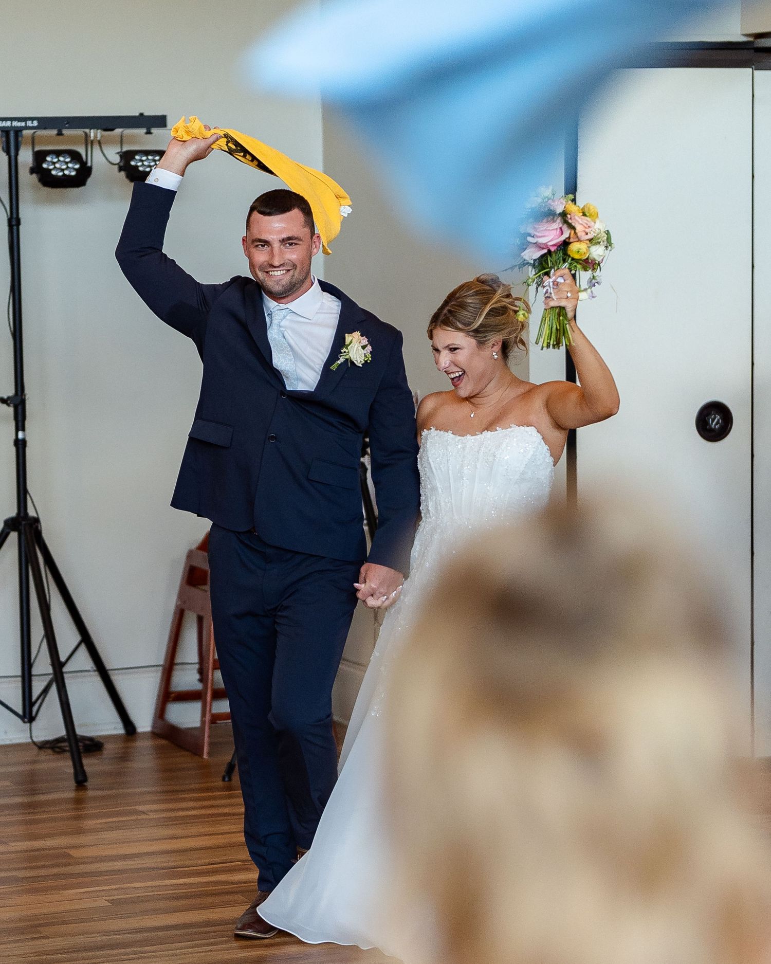 Bride and groom make grand entrance at wedding reception, groom waving yellow towel joyfully.