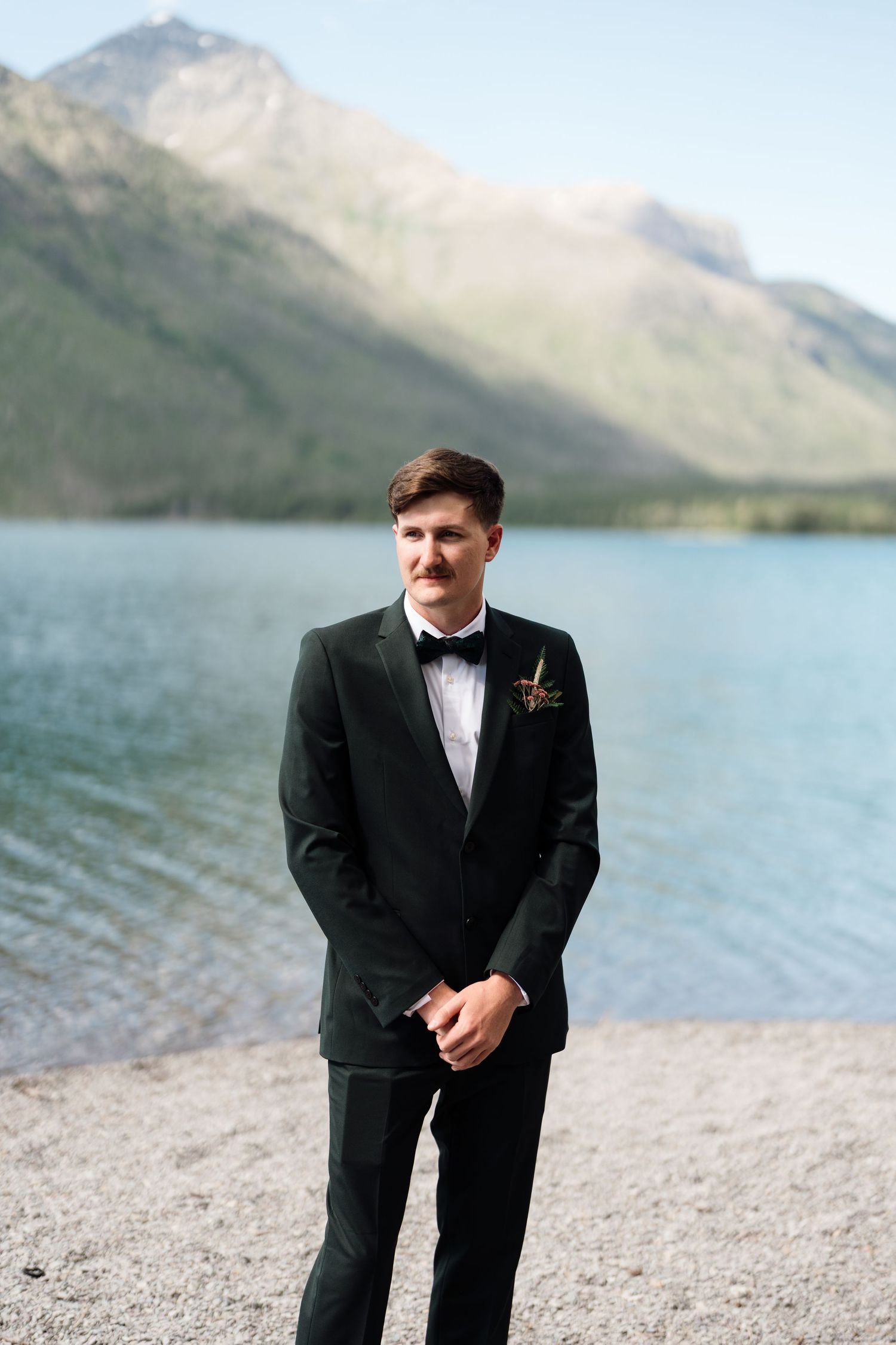Groom in black tuxedo with boutonniere stands by a mountain lake on a sunny wedding day.