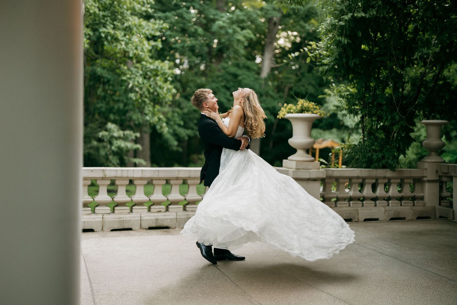 Groom twirling bride in white gown on elegant stone terrace surrounded by lush green trees at outdoor wedding.