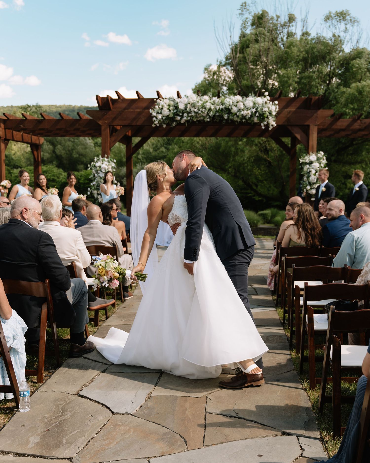Bride and groom share first kiss as newlyweds walking down outdoor wedding aisle lined with guests under floral pergola.