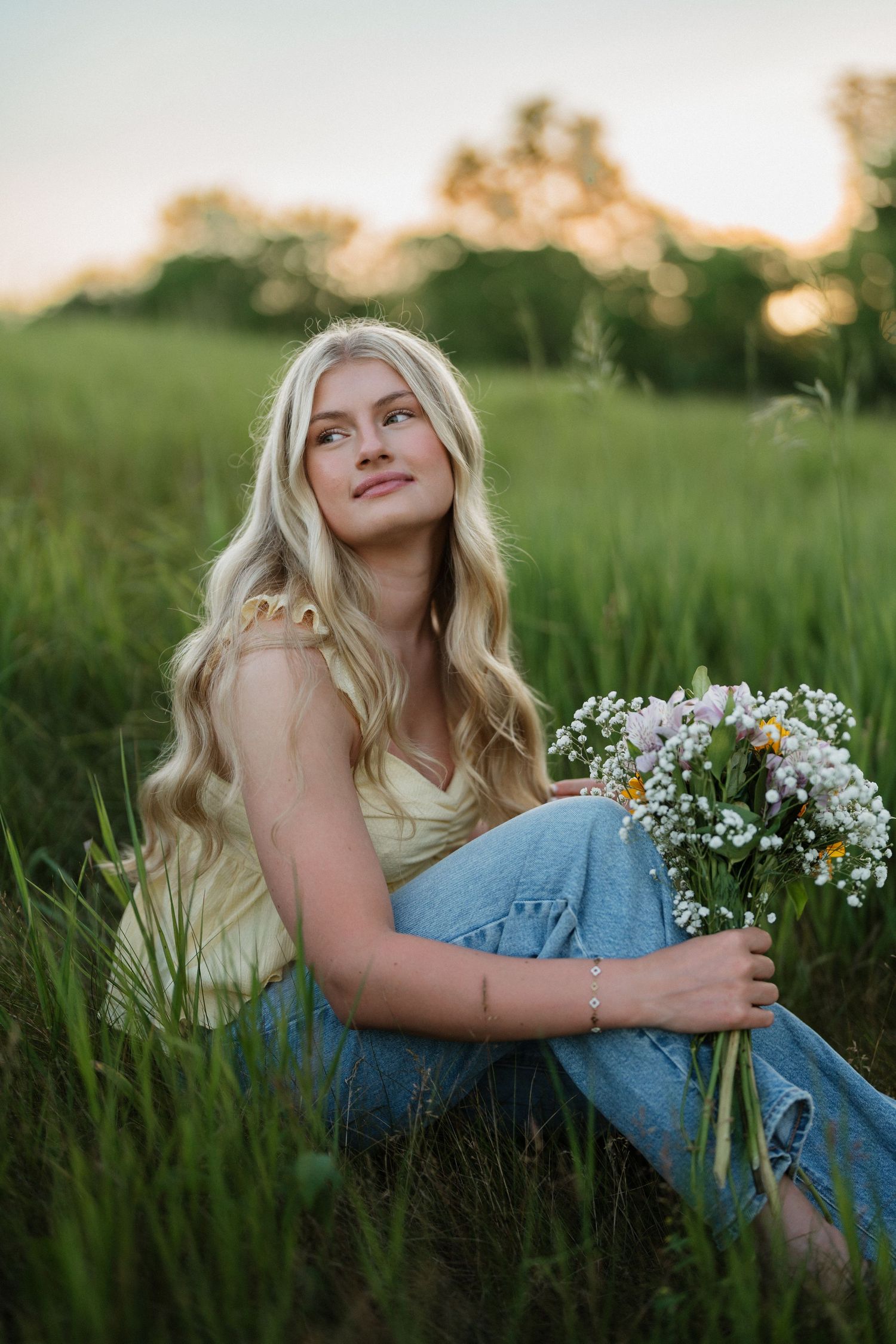 Blonde woman sitting in green field holding wildflowers, wearing yellow top and jeans at sunset.