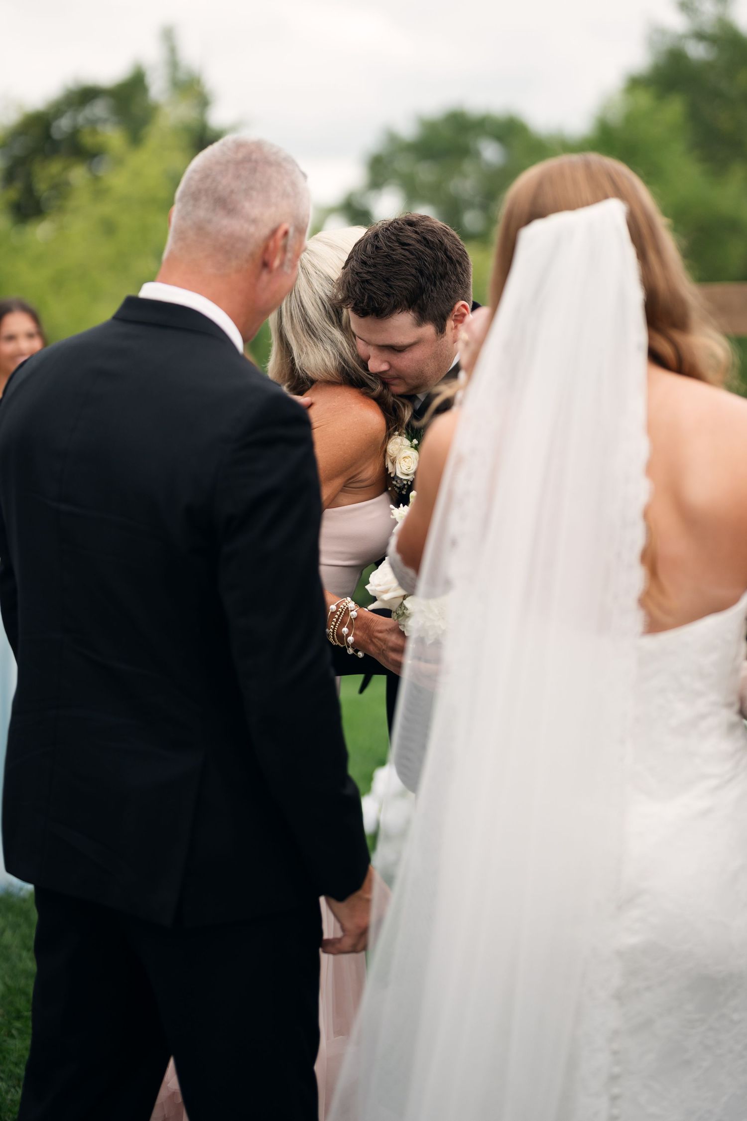Groom sharing an emotional moment with his mother during an outdoor wedding ceremony while bride watches.