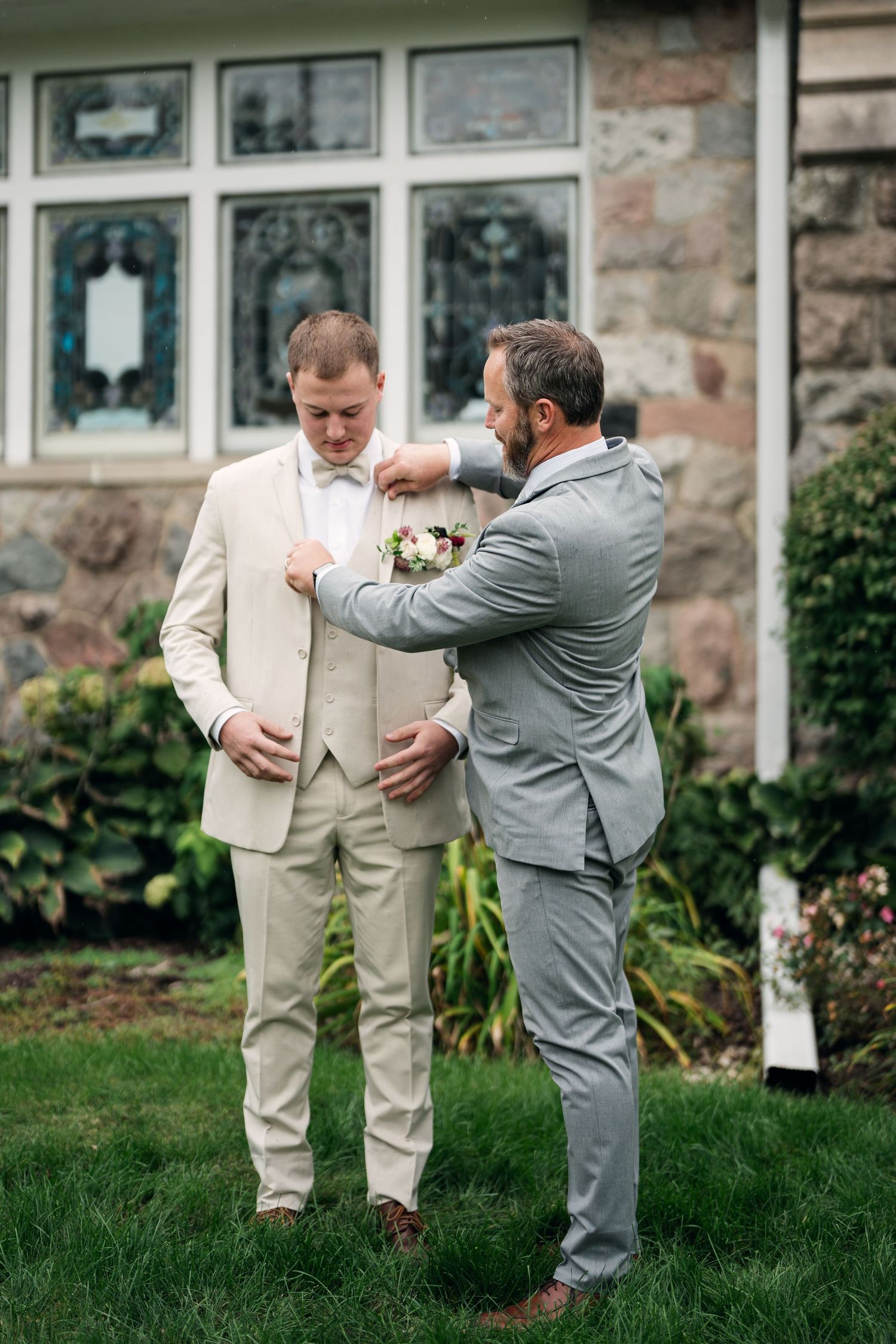 Groomsman pinning boutonniere on groom in cream suit outside stone church.