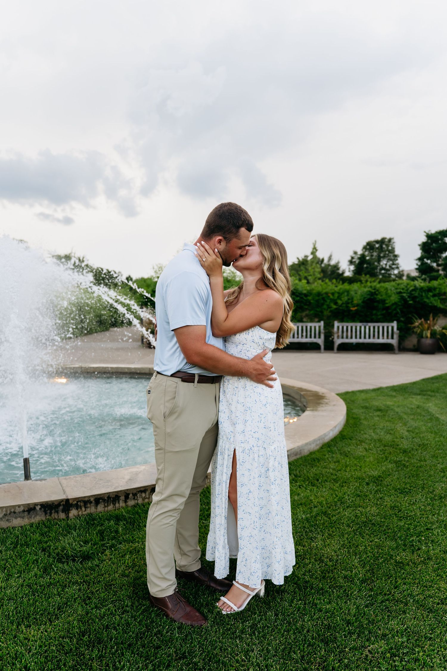 A couple shares a romantic moment by a fountain in a garden setting with water features and green lawn.