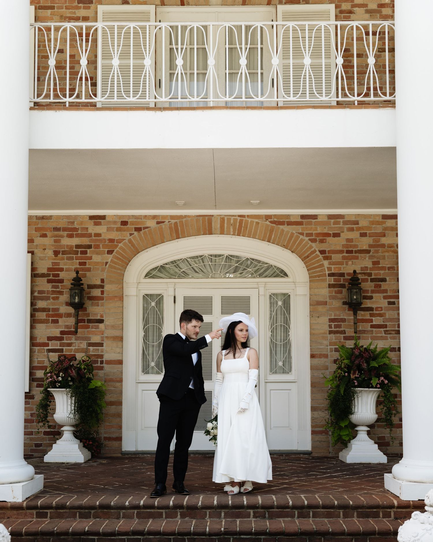 A bride and groom share a quiet moment on the brick porch of a white-columned building with elegant ironwork details.