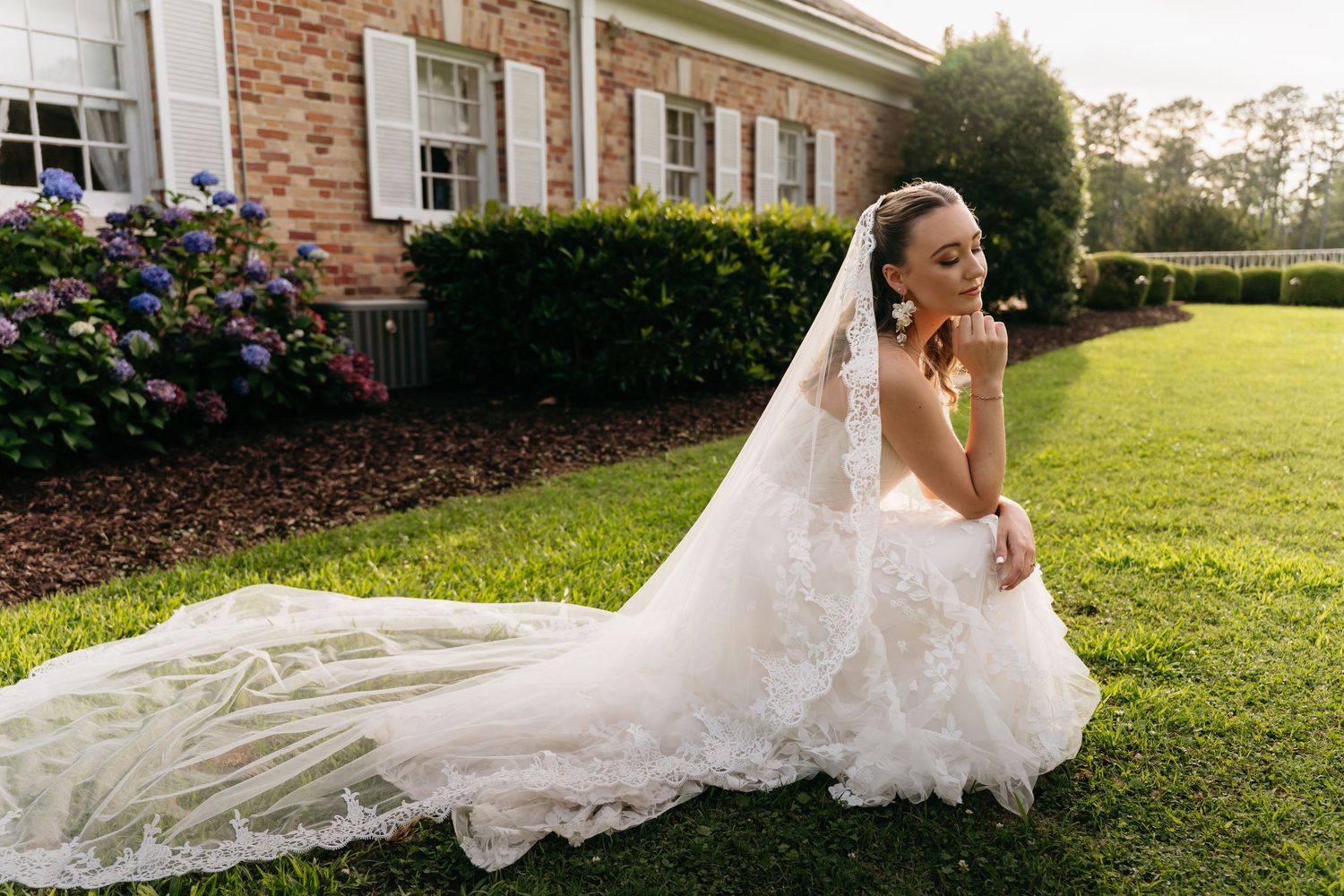 A flowing white wedding dress with a long train spreads gracefully across a green lawn outside a brick building.