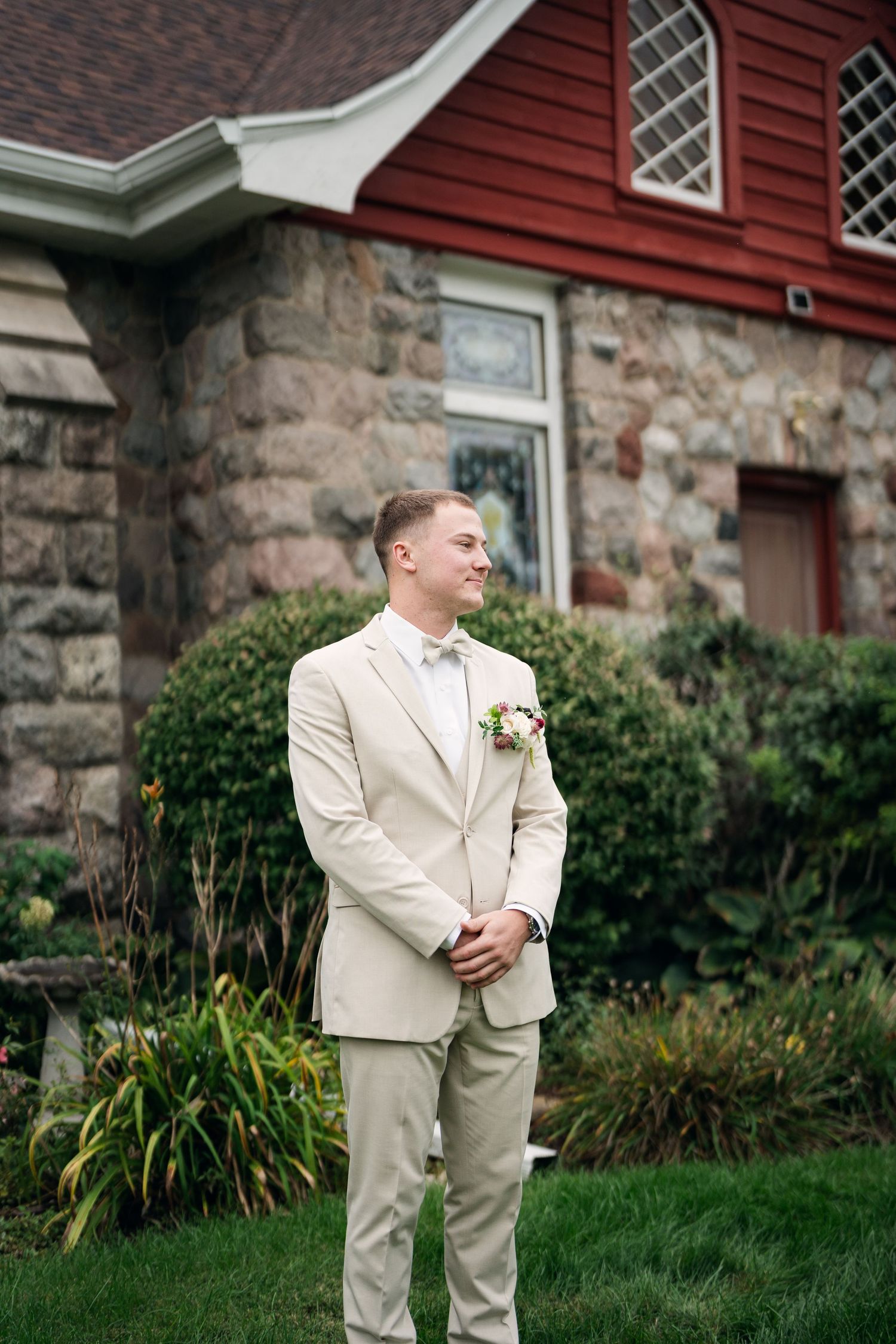 Groom in beige suit with boutonniere stands smiling outdoors before stone and red wood building with lush greenery.