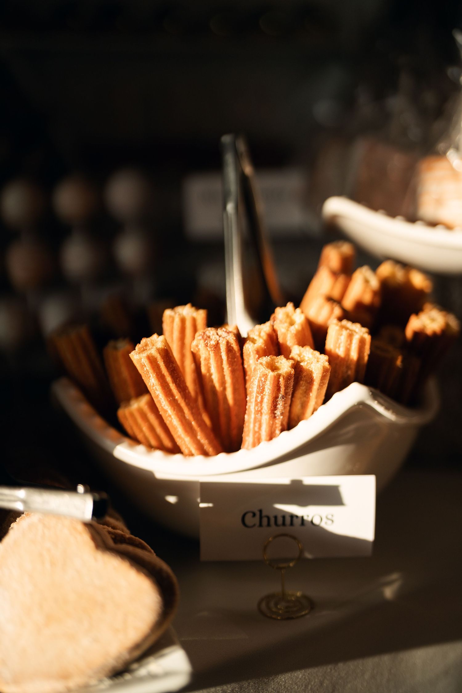Golden churros displayed in a white ceramic bowl with tongs and a label sign at a buffet dessert station.