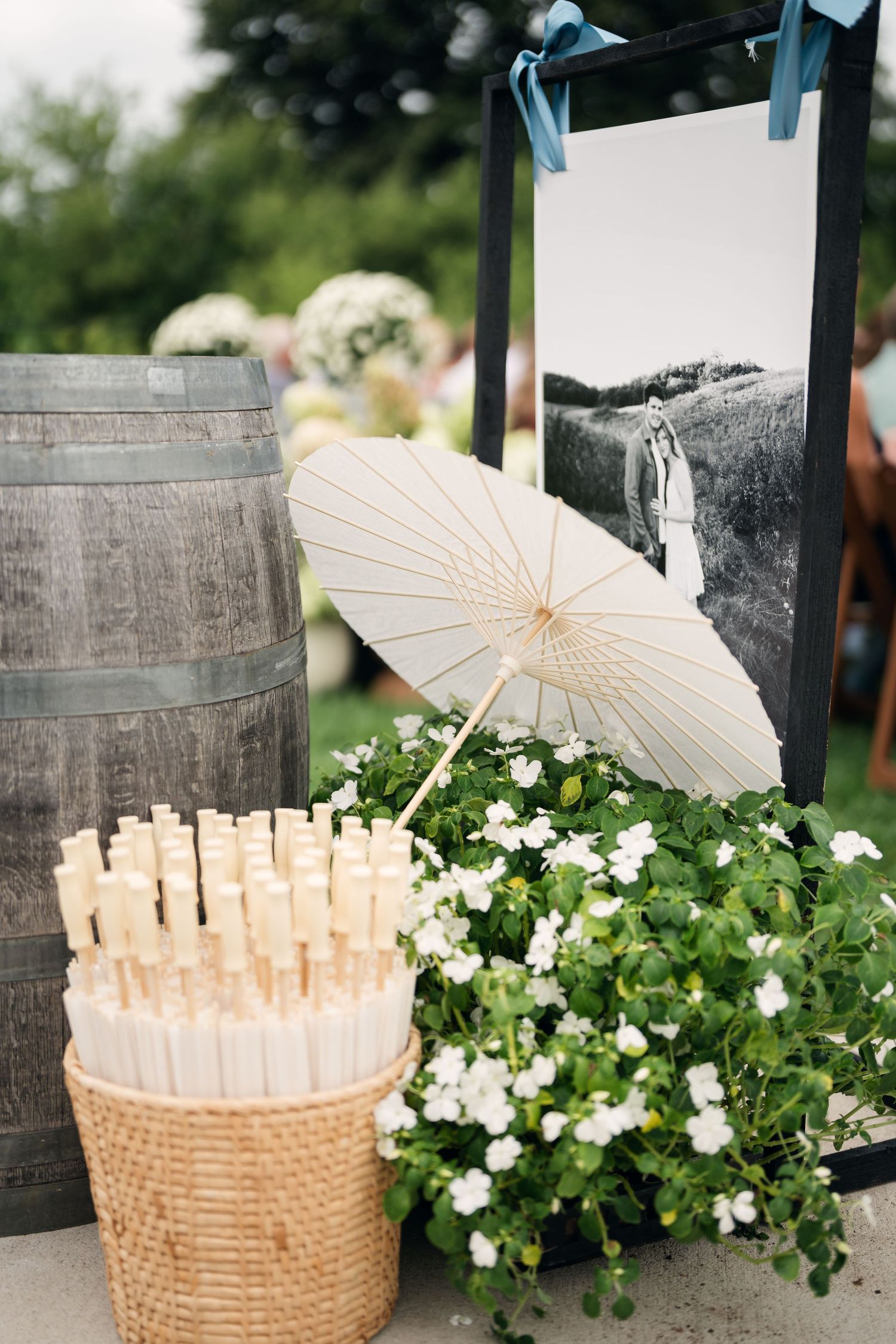 Wedding ceremony decor with parasols in wicker basket, wine barrel, white flowers, and black and white photo display.