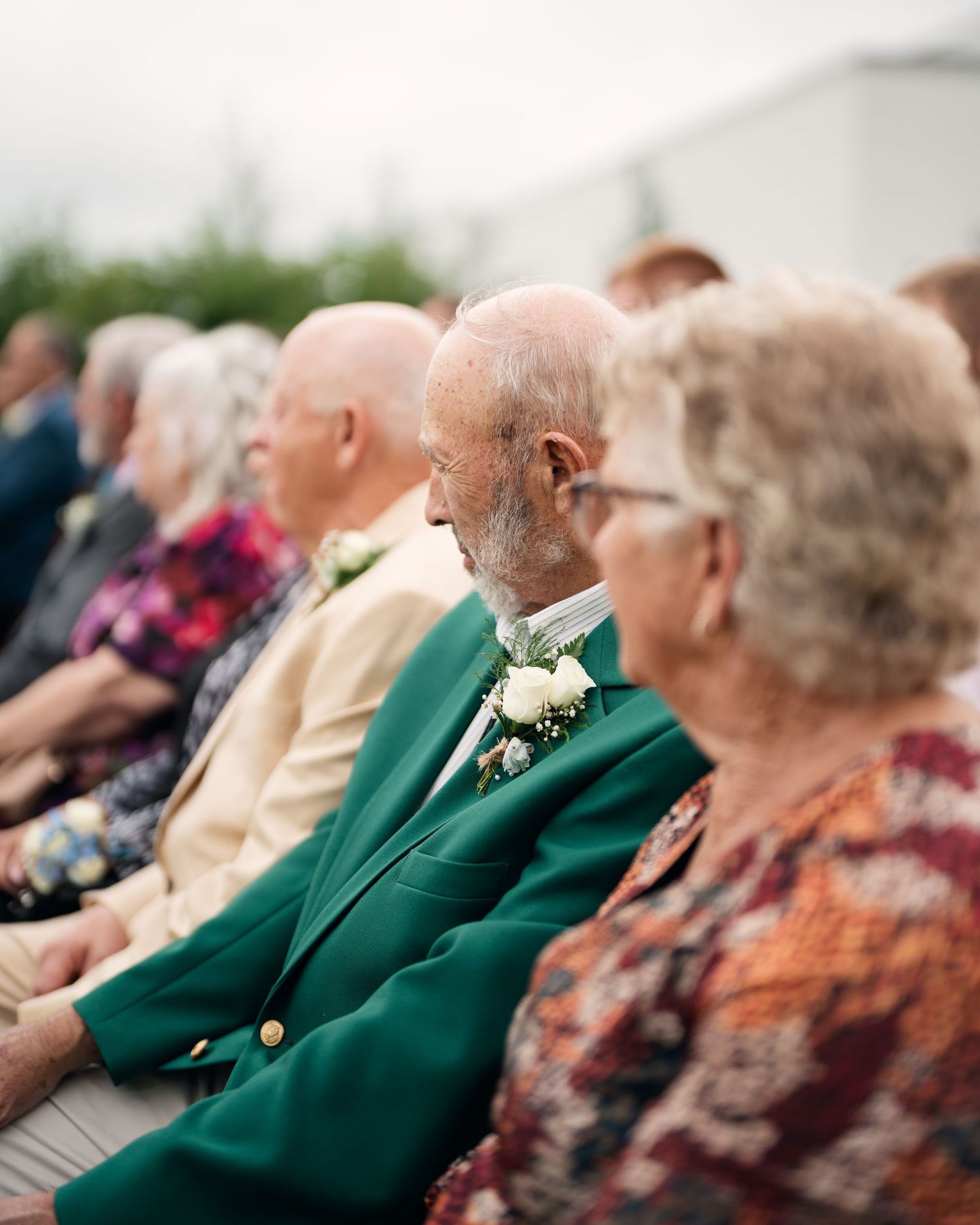 Elderly wedding guests seated outdoors, man in green jacket wearing white boutonniere at ceremony.