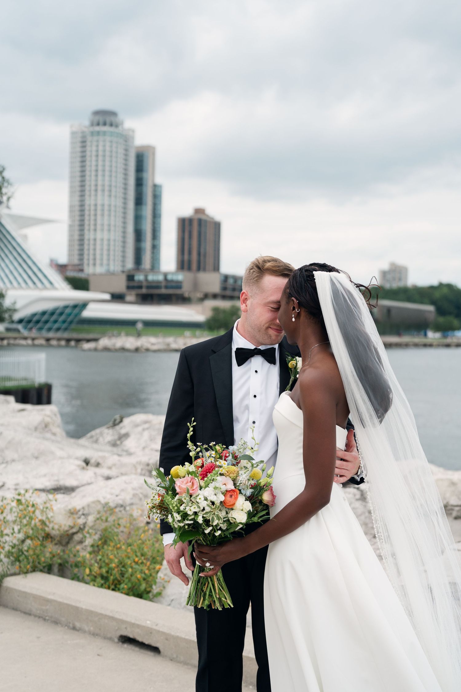 Interracial couple sharing a kiss on their wedding day by a waterfront, bride holding colorful bouquet, city skyline behind them.