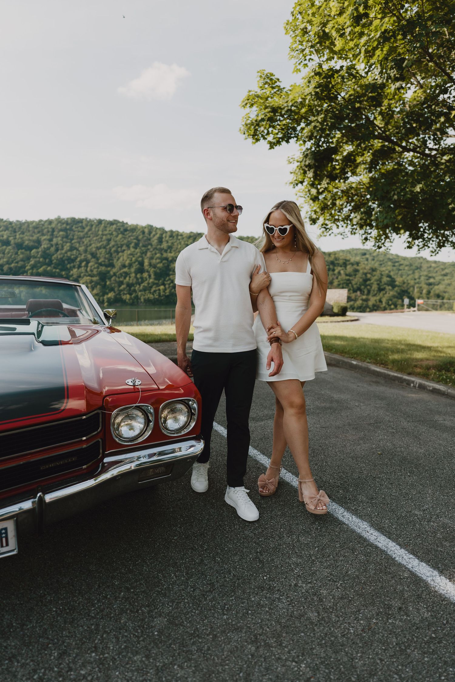 Couple posing with vintage red car against mountain landscape backdrop.