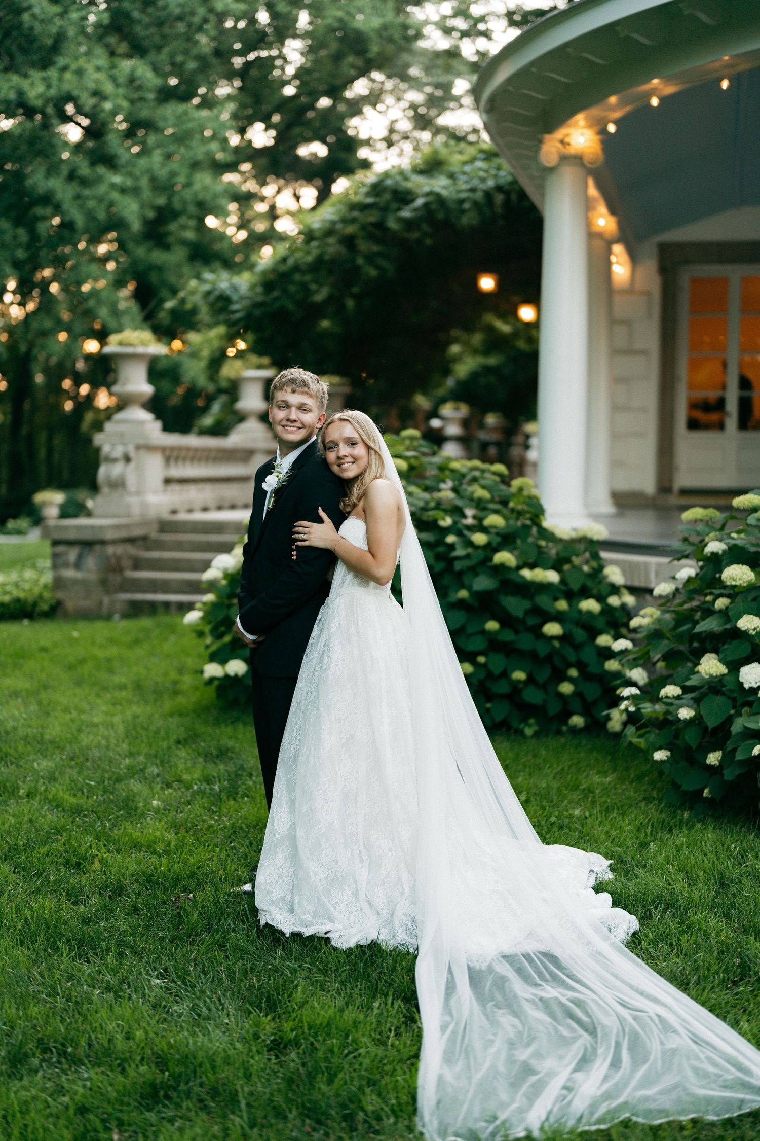 A smiling bride in a flowing white gown with veil embraces a groom in a black tuxedo on a lush green lawn.