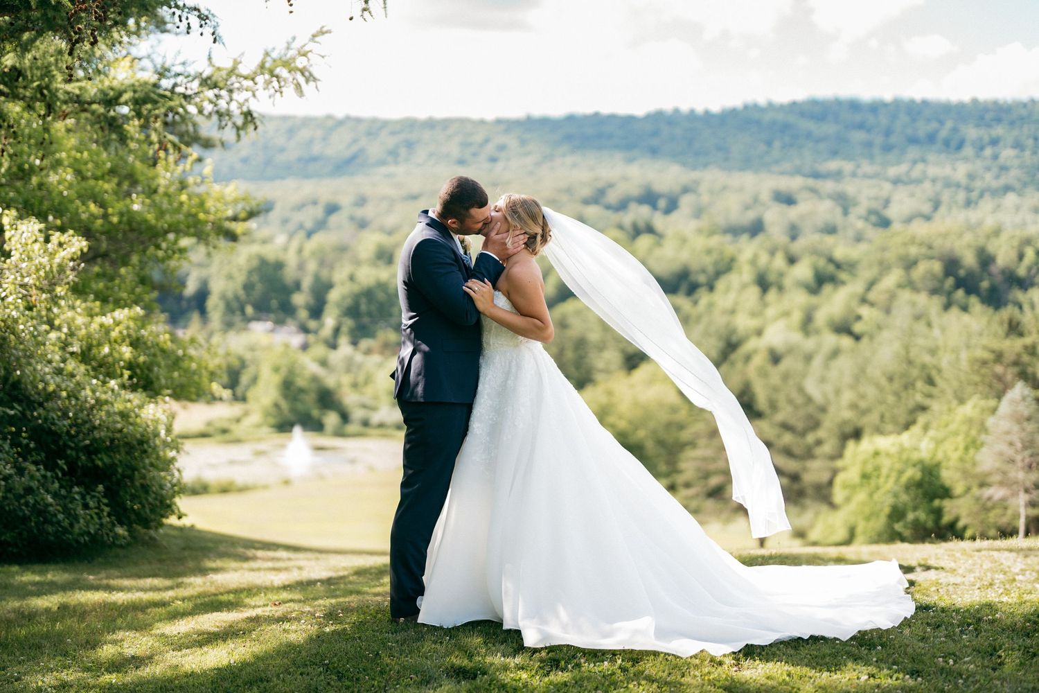 Bride and groom share a romantic kiss outdoors on their wedding day with lush green hills in the background.