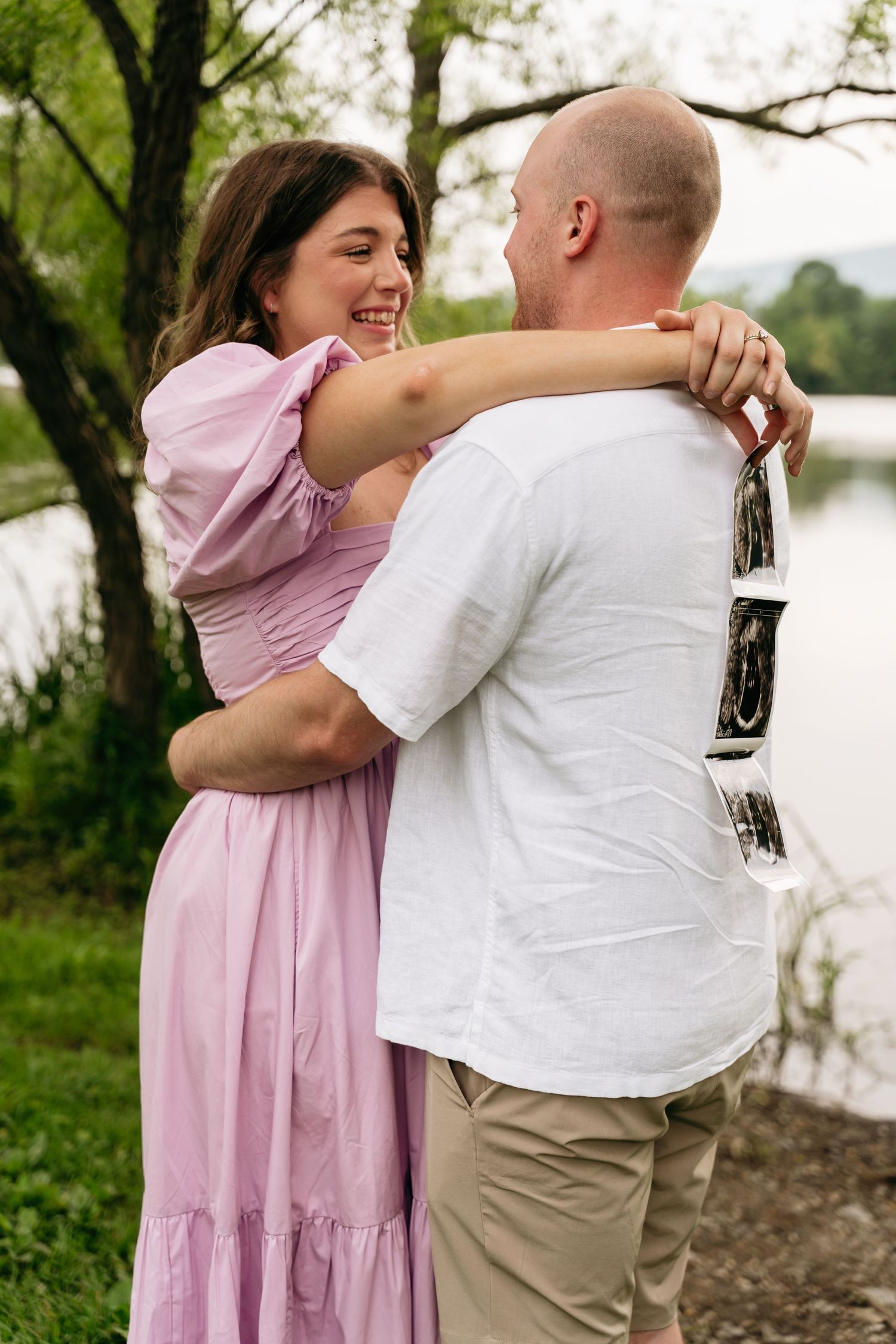 A couple embraces outdoors by a lake, with the woman wearing a pink dress and the man in a white shirt and khakis.