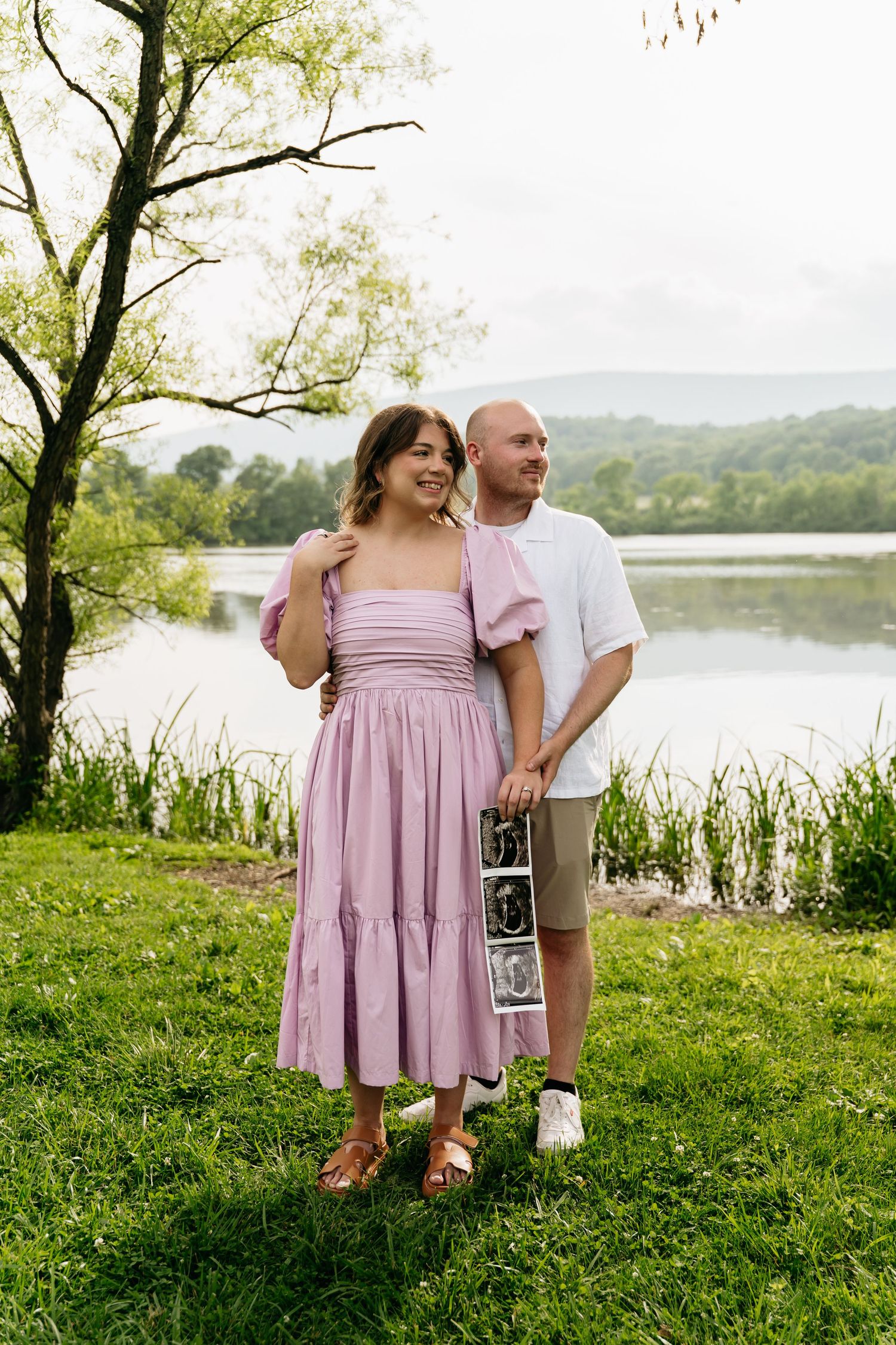 Romantic couple stands by a serene lake on grassy shore while holding a sonogram photo during their pregnancy announcement.