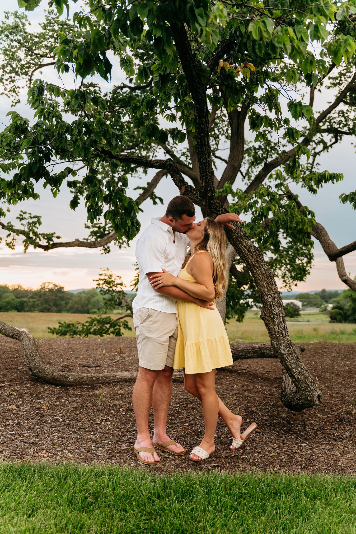 A romantic couple in light summer attire shares tender moments under a leafy tree at sunset.