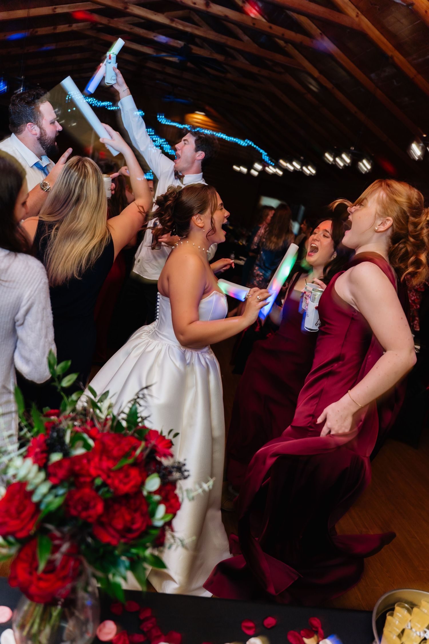 Bride and bridesmaids in burgundy dresses dancing joyfully with glowsticks at a wedding reception.